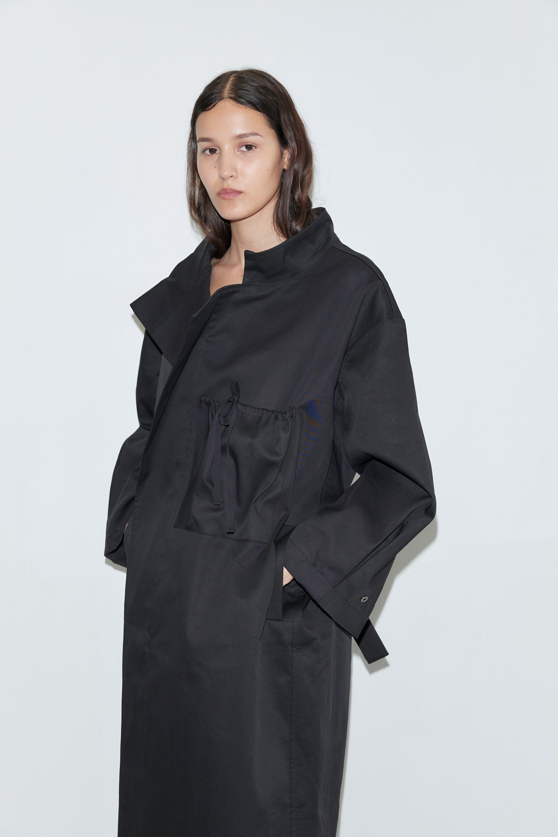 A woman with long dark hair models the Longline Jacket - Black, featuring large pockets, as she stands against a plain white background and looks neutrally at the camera.