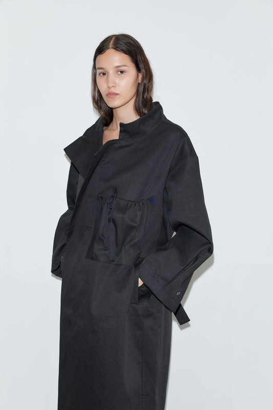A woman with long dark hair models the Longline Jacket - Black, featuring large pockets, as she stands against a plain white background and looks neutrally at the camera.