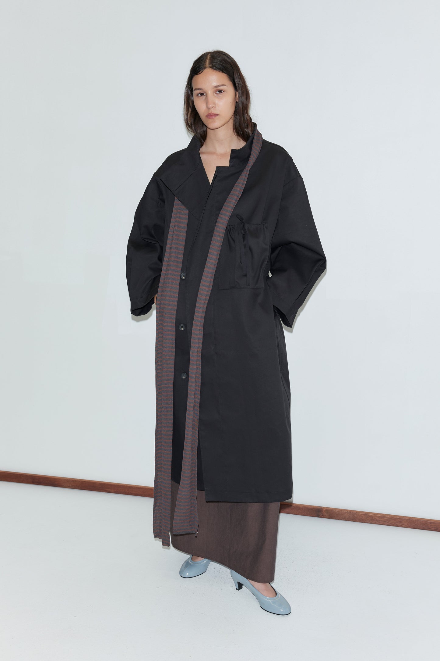 A woman stands by a plain white wall, wearing a long black overcoat with wide sleeves, brown trim, and pockets. She layers her outfit with the Knit Scarf - Brown Grey Stripe and light blue shoes, her long dark hair worn loose.