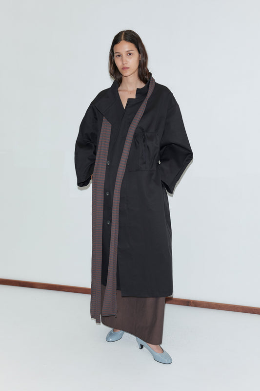 A woman stands by a plain white wall, wearing a long black overcoat with wide sleeves, brown trim, and pockets. She layers her outfit with the Knit Scarf - Brown Grey Stripe and light blue shoes, her long dark hair worn loose.