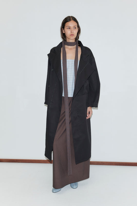 A woman with loose brown hair and a neutral expression stands against a white background wearing a Longline Jacket - Black, a light blue top, long brown skirt, matching scarf, and light blue shoes.