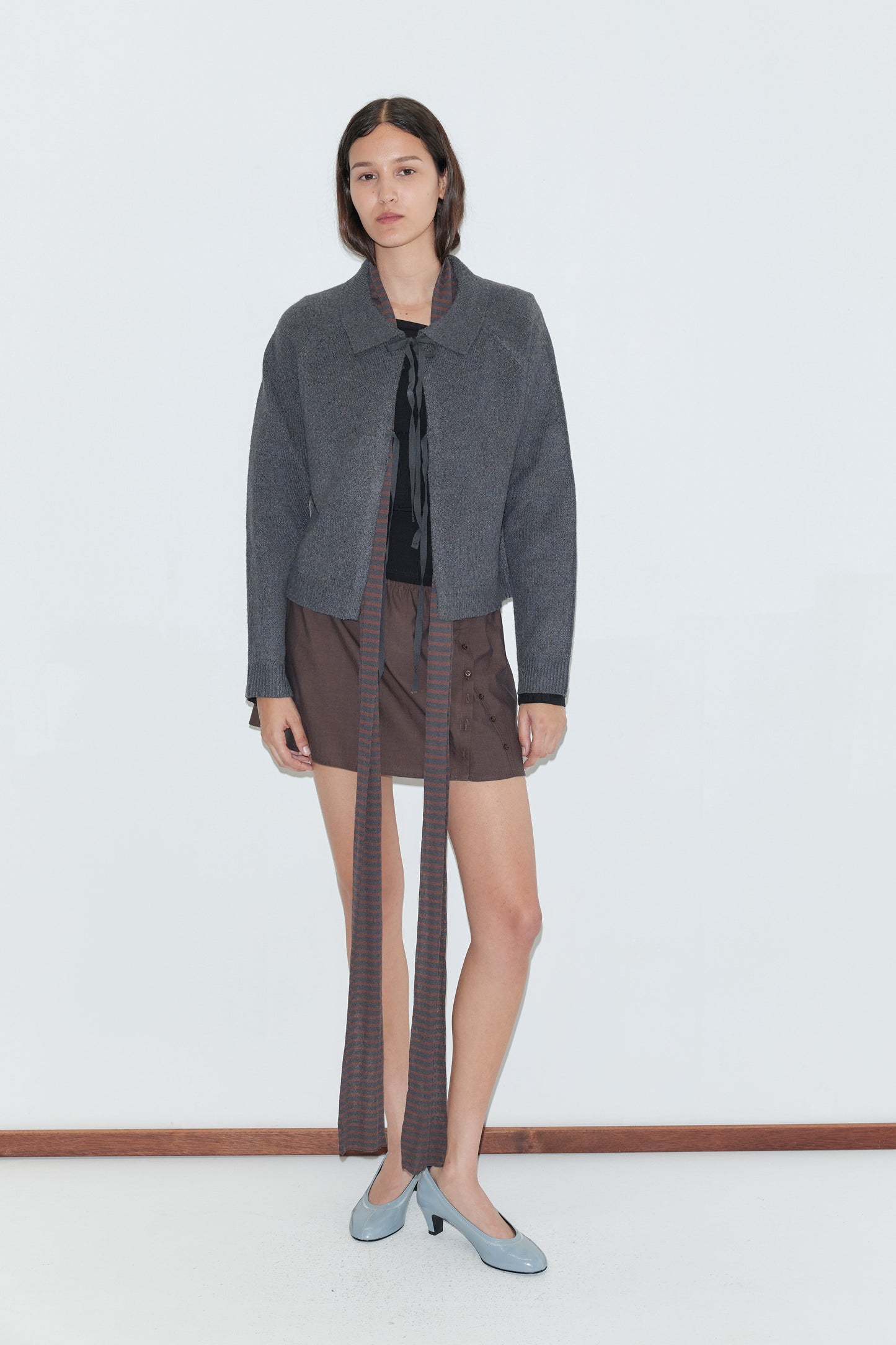 A woman with straight dark hair and a neutral expression stands against a white background wearing a Deiji Studios Box Cardigan - Charcoal, paired with a brown mini skirt, black top, and light blue shoes.