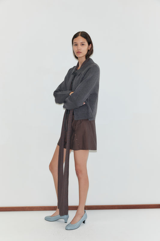 A woman with long dark hair and a neutral expression stands against a white background, arms crossed, wearing the Box Cardigan - Charcoal with a brown skirt and light blue heeled shoes.