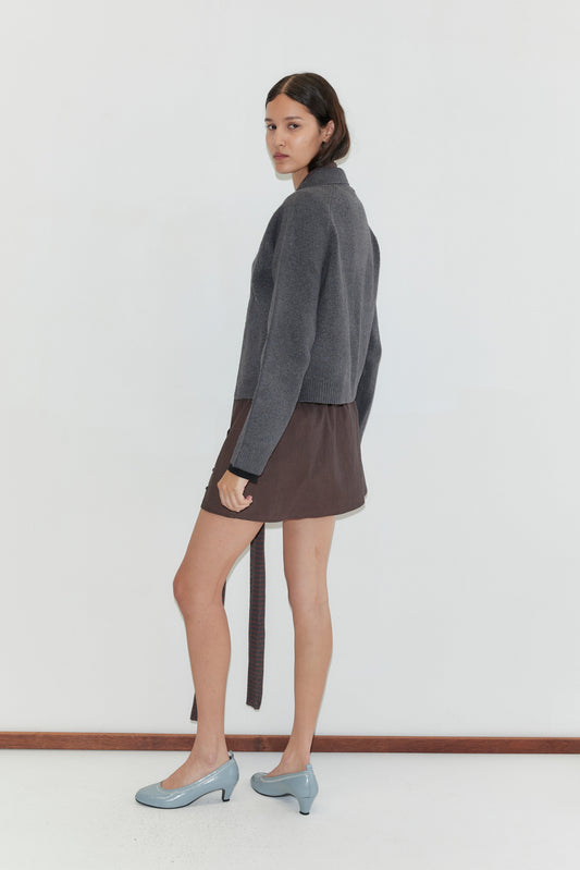 A woman with dark hair stands sideways against a white background, wearing the Box Cardigan in Charcoal by Deiji Studios, a brown skirt with a long belt, and light blue low-heeled shoes. She looks over her shoulder at the camera.