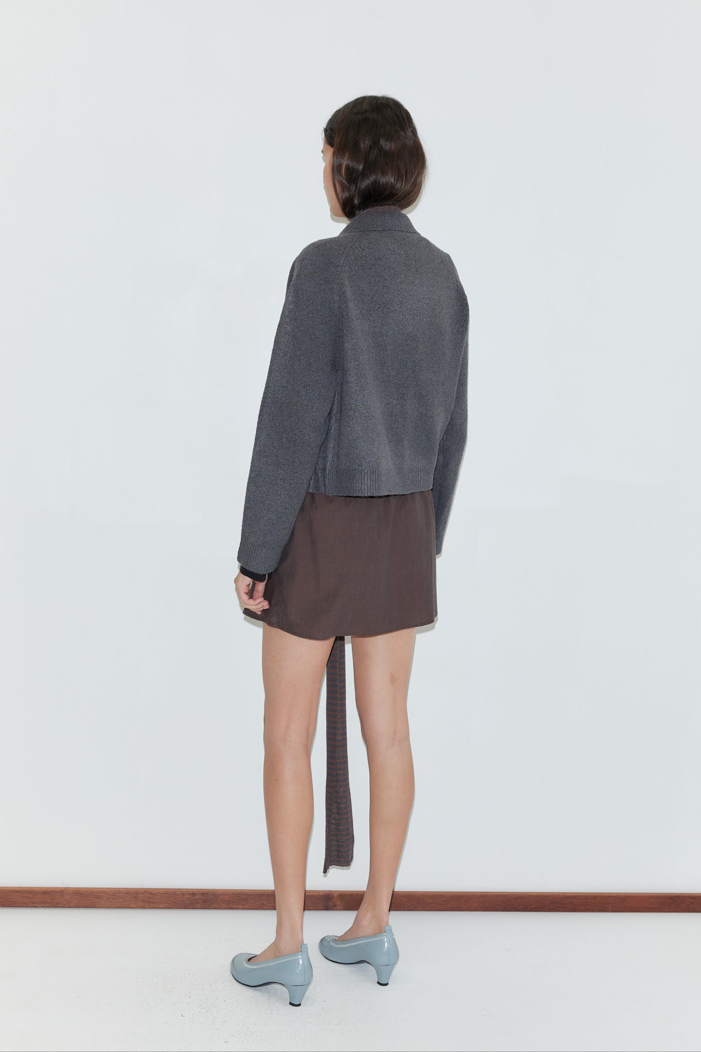 A person stands facing away, wearing the Box Cardigan - Charcoal with a brown asymmetrical skirt and light blue heeled shoes, against a plain white background.