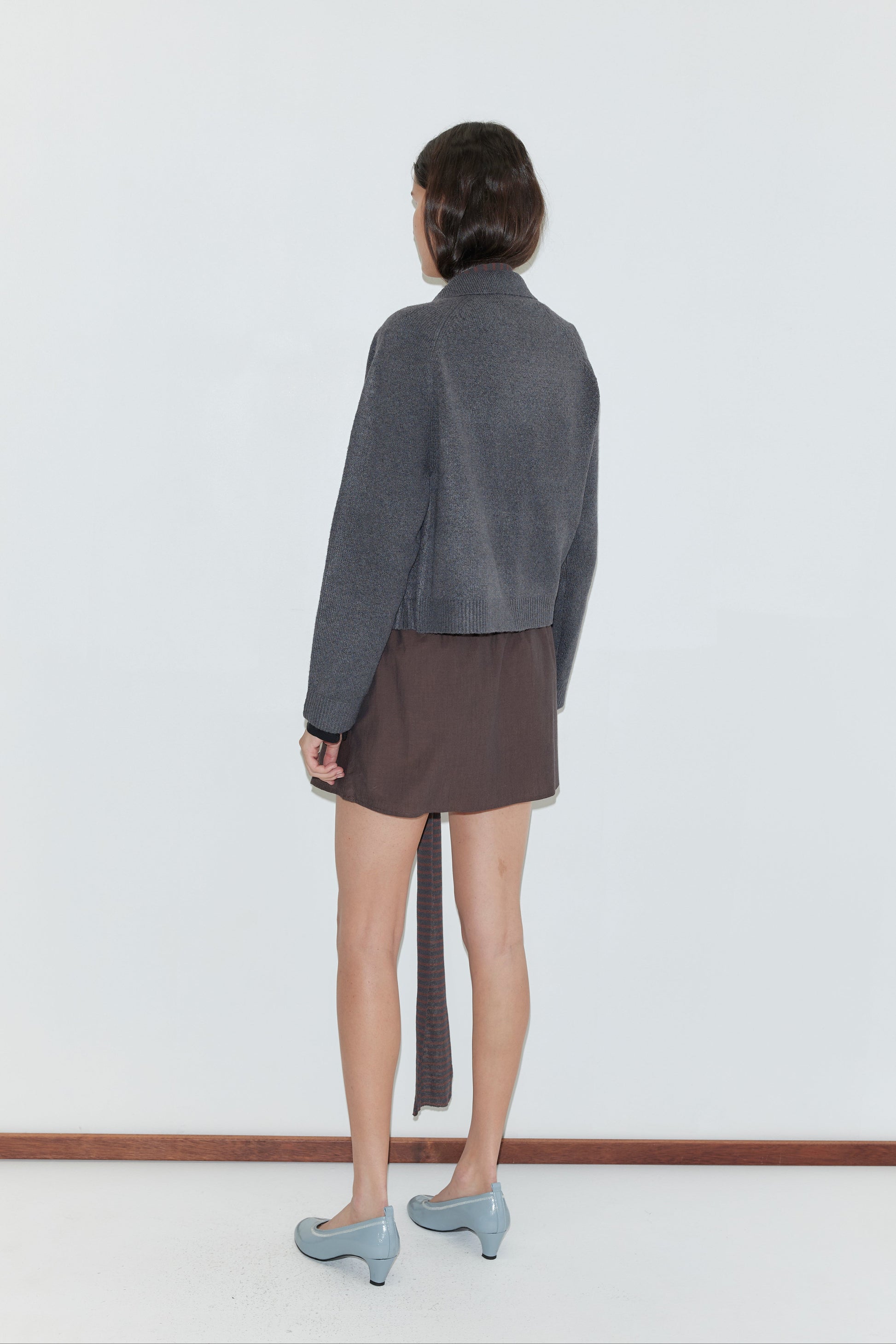 A person stands facing away, wearing the Box Cardigan - Charcoal with a brown asymmetrical skirt and light blue heeled shoes, against a plain white background.