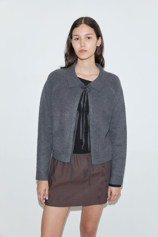 A young woman with long dark hair stands against a plain white background, wearing the Box Cardigan in Charcoal over a black top with ties and a brown skirt with side buttons.