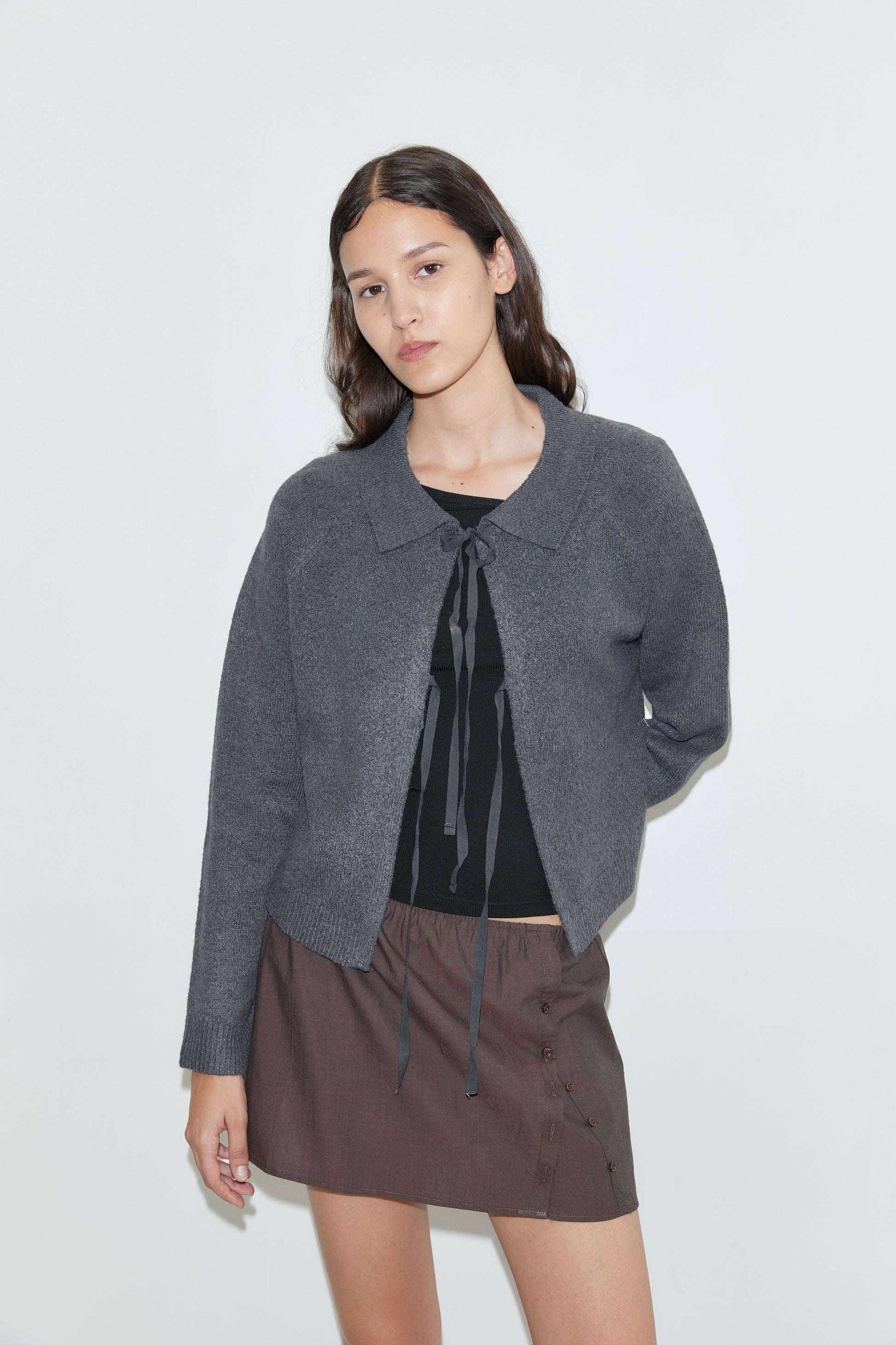 A woman with long dark hair wears the Box Cardigan in Charcoal over a black tie-detail top and a brown buttoned skirt, standing against a plain white background.