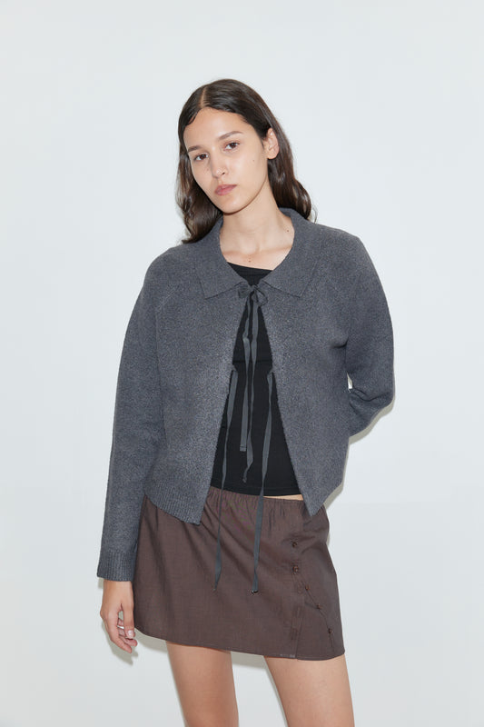A woman with long dark hair wears the Box Cardigan in Charcoal over a black tie-detail top and a brown buttoned skirt, standing against a plain white background.
