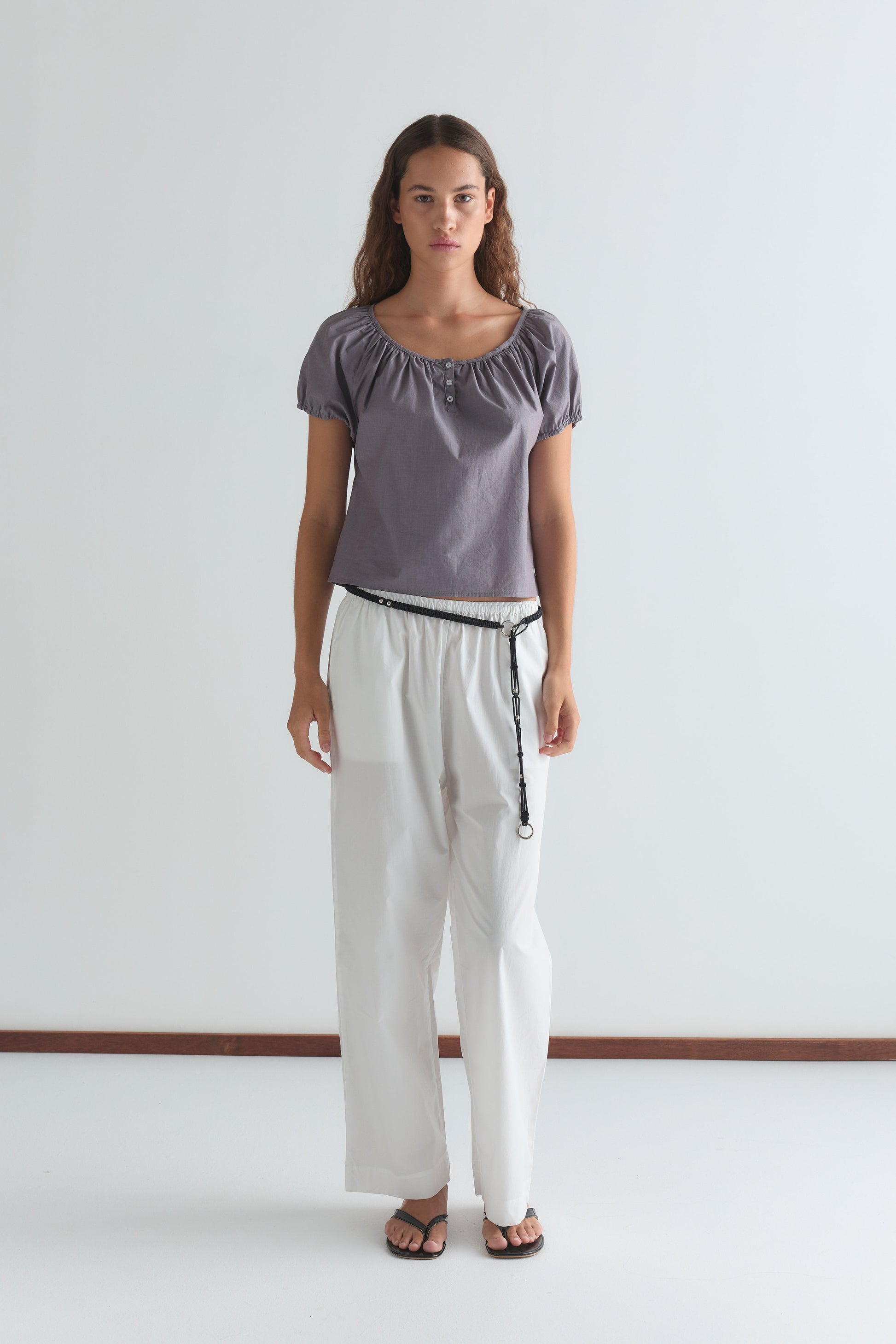 A woman with long, wavy brown hair and a neutral expression stands against a white background wearing the Poplin T Shirt - Currant by Deiji Studios, paired with loose white pants, a black belt, and black sandals.