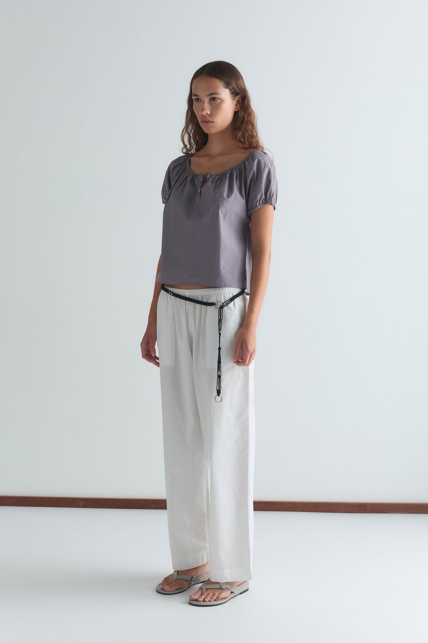 A woman with long, wavy brown hair stands in a minimal room wearing the Deiji Studios Poplin T Shirt - Currant, paired with loose white pants and sandals. She faces slightly to the side with a neutral expression.