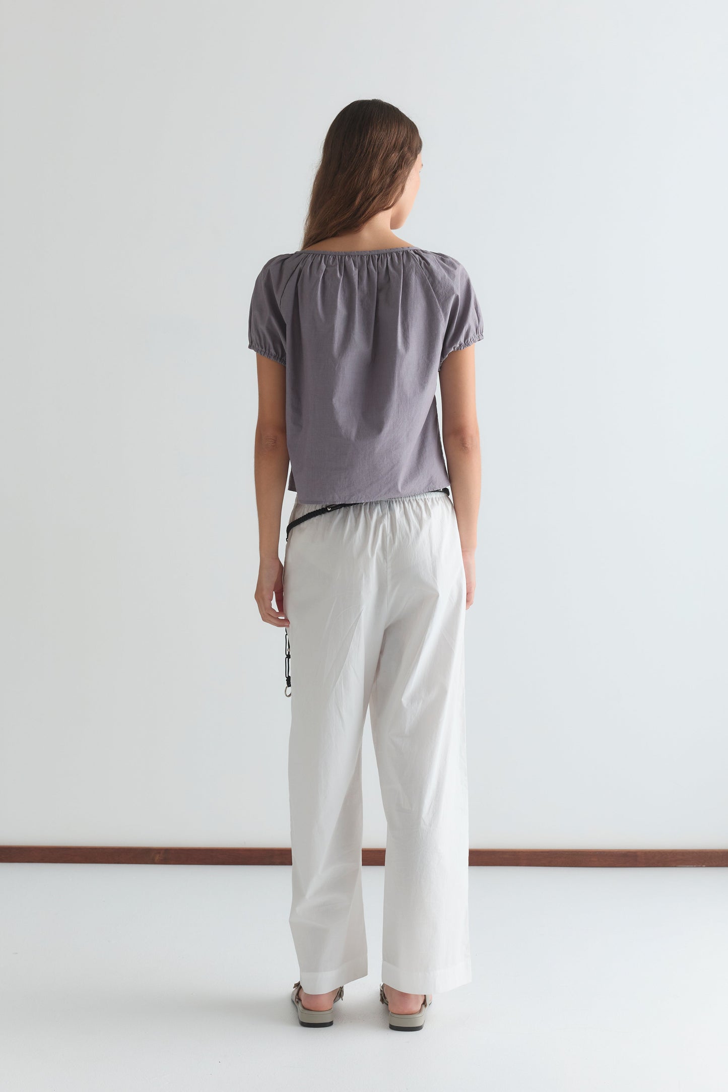 A woman faces away from the camera wearing the Poplin T Shirt - Currant, featuring a loose, cropped relaxed silhouette, paired with white wide-leg pants and sandals against a plain white background.