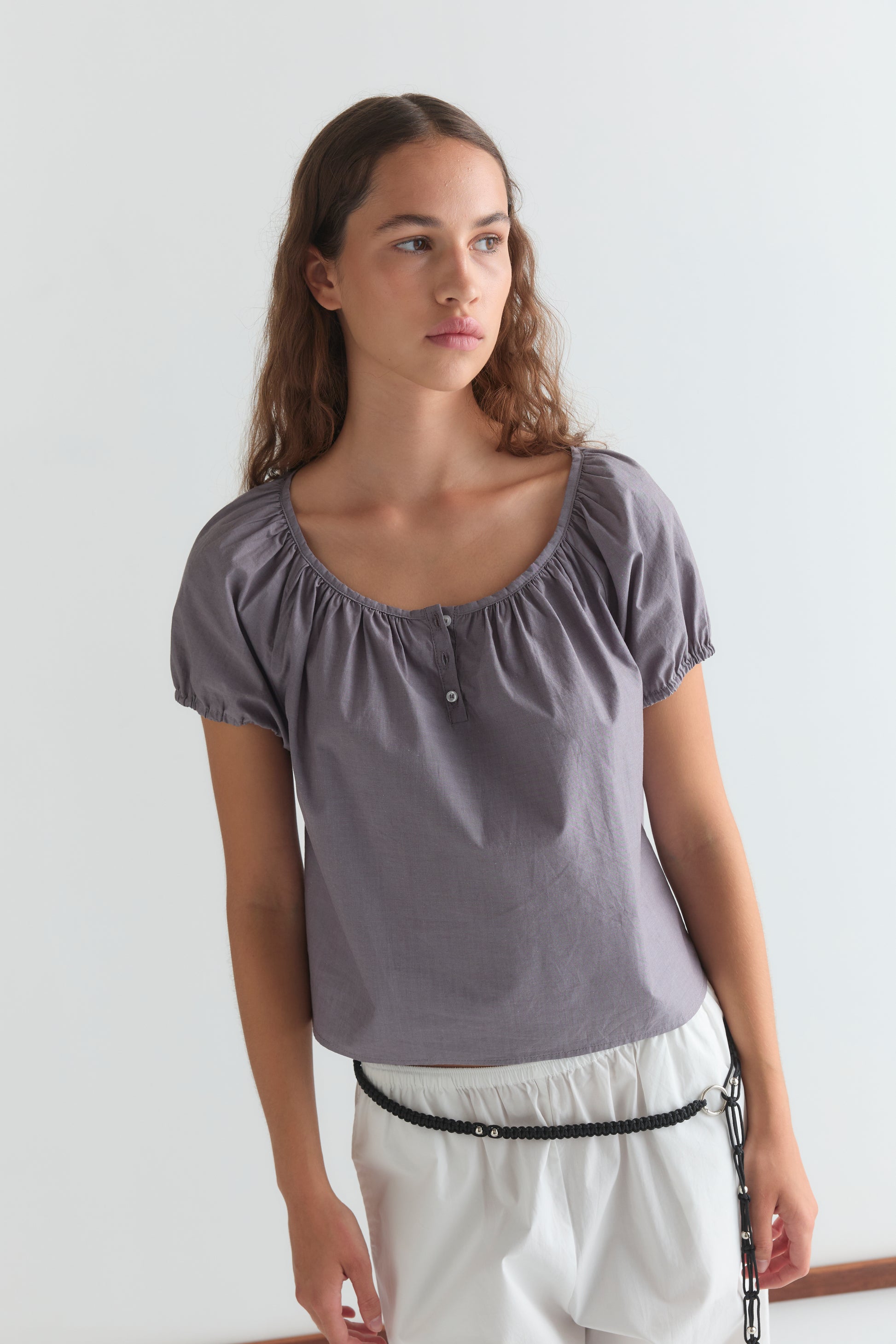 A woman with long brown hair wears the Deiji Studios Poplin T Shirt - Currant in a cropped relaxed fit and white shorts with a black braided belt, standing against a plain white background and looking to the side.