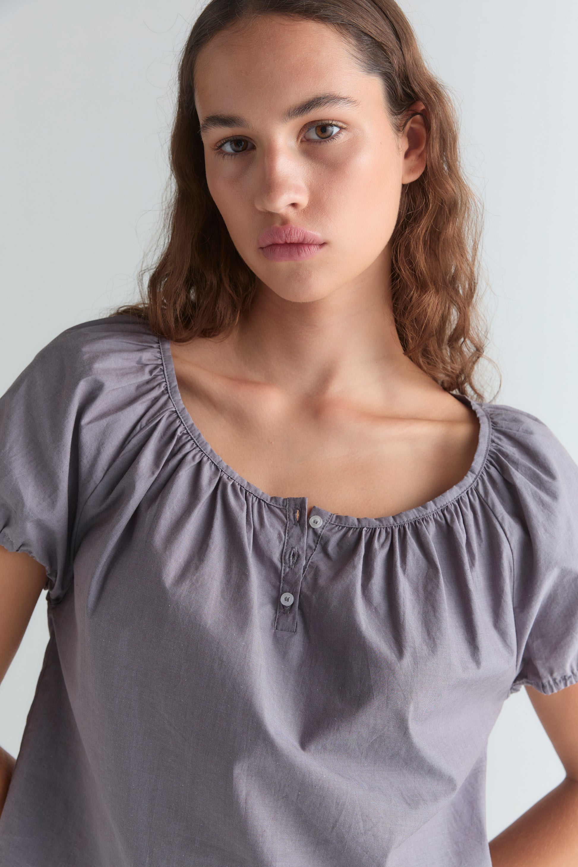 A young woman with long brown hair wears the Poplin T Shirt - Currant by Deiji Studios, featuring a cropped relaxed fit and gathered neckline. She faces the camera with a neutral look against a light background.