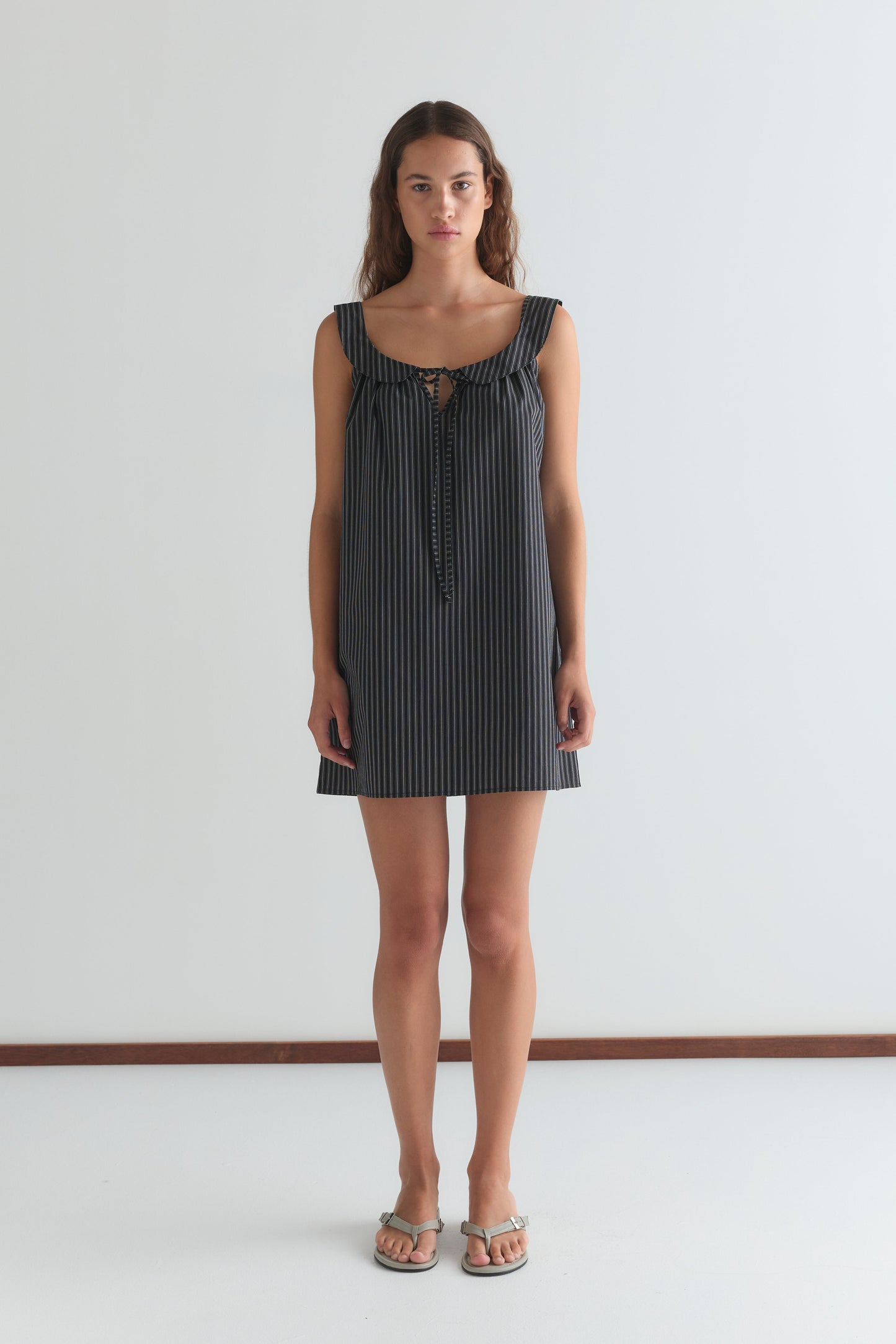 A woman stands against a white wall, wearing the Rounded Neck Dress - Mono stripe by Deiji Studios—a sleeveless, navy and white striped mini dress with front tie detail—paired with silver sandals. She faces forward with a neutral expression.