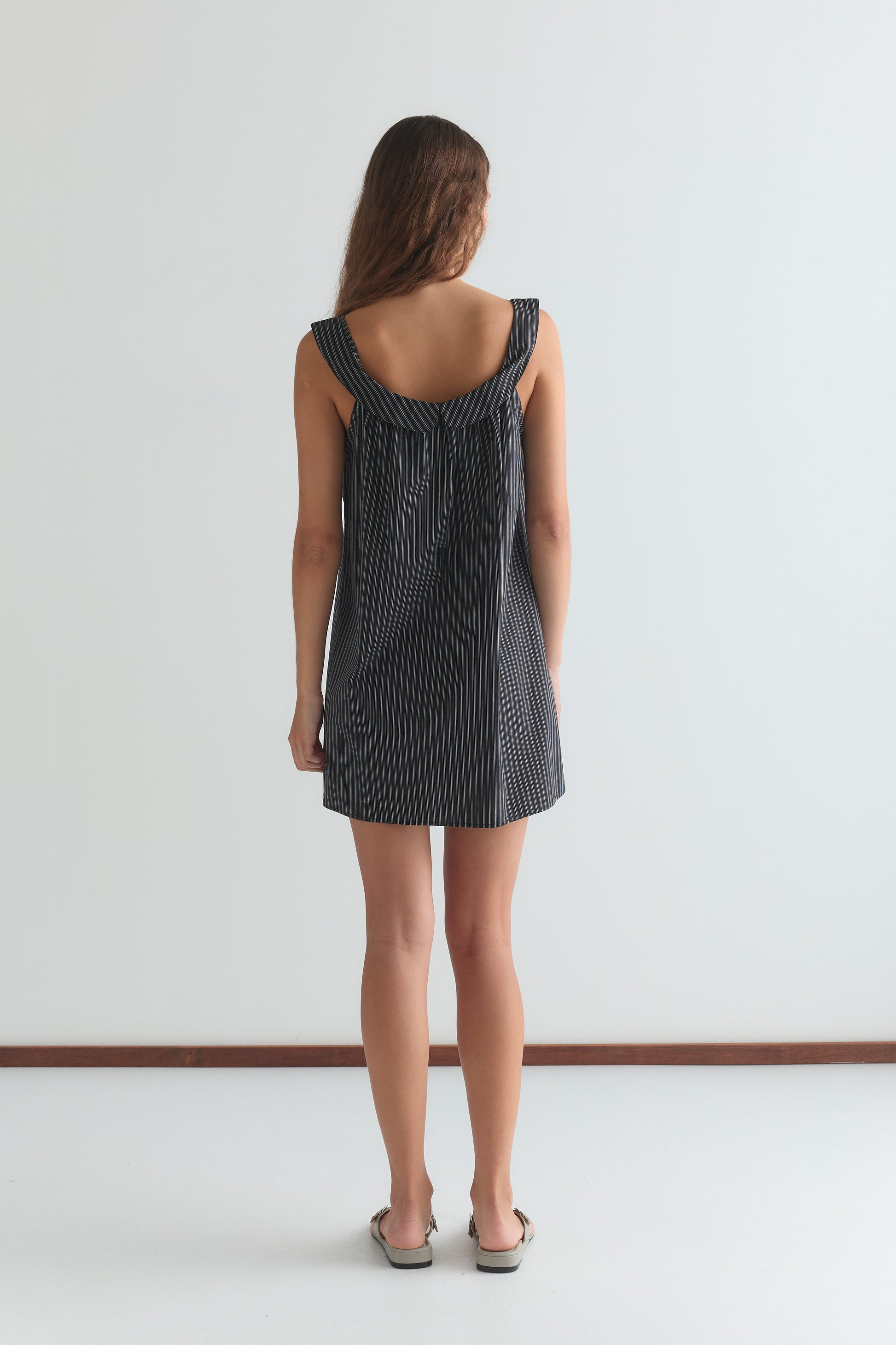 A woman with long brown hair stands facing away in a minimalist room, wearing the Rounded Neck Dress - Mono stripe by Deiji Studios and sandals. White walls and a wooden floor trim create a simple backdrop.
