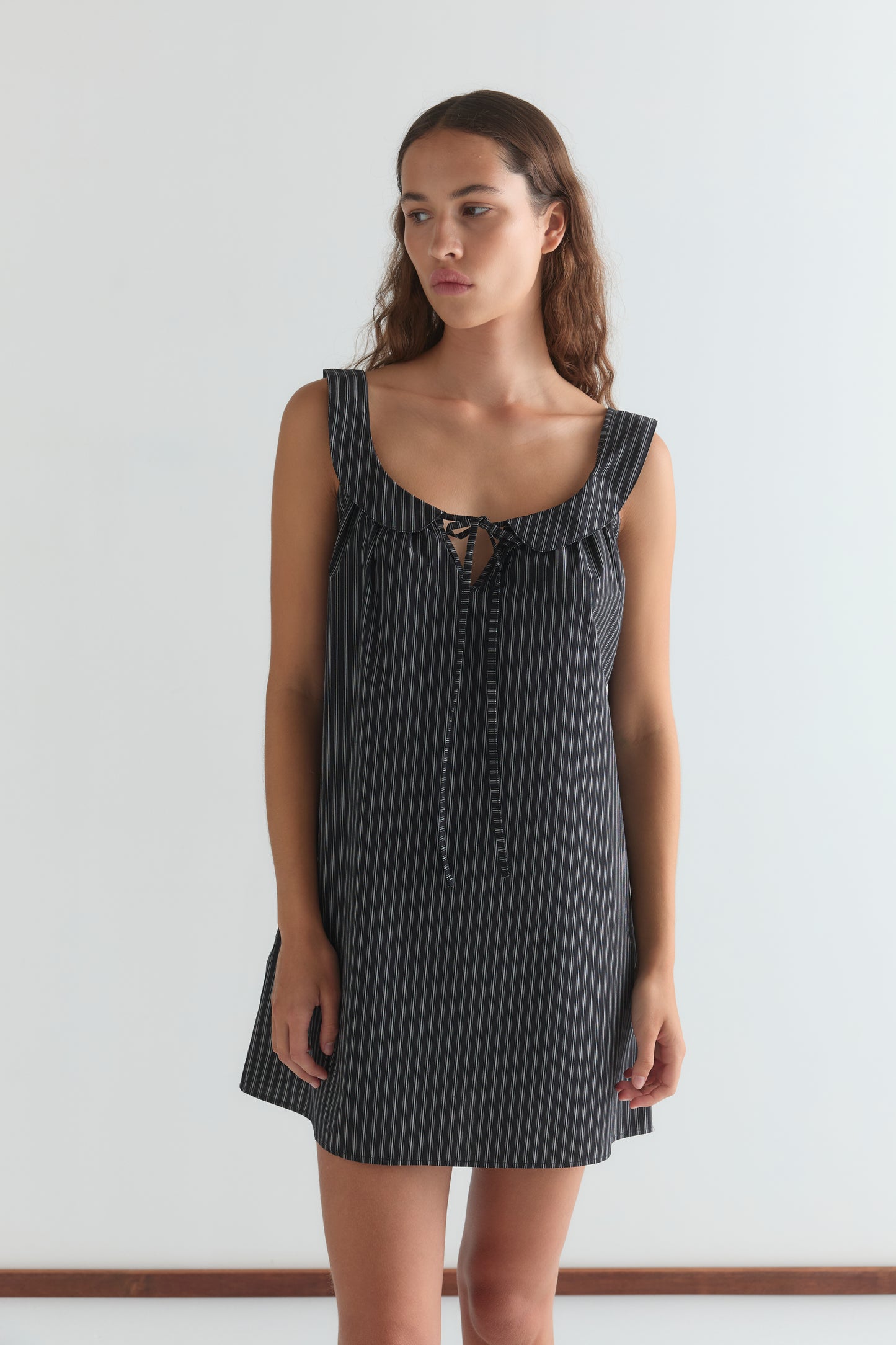 A woman with long brown hair wears the Rounded Neck Dress - Mono stripe, a sleeveless short black dress with white vertical stripes and a tie at the neckline, standing against a plain white background.