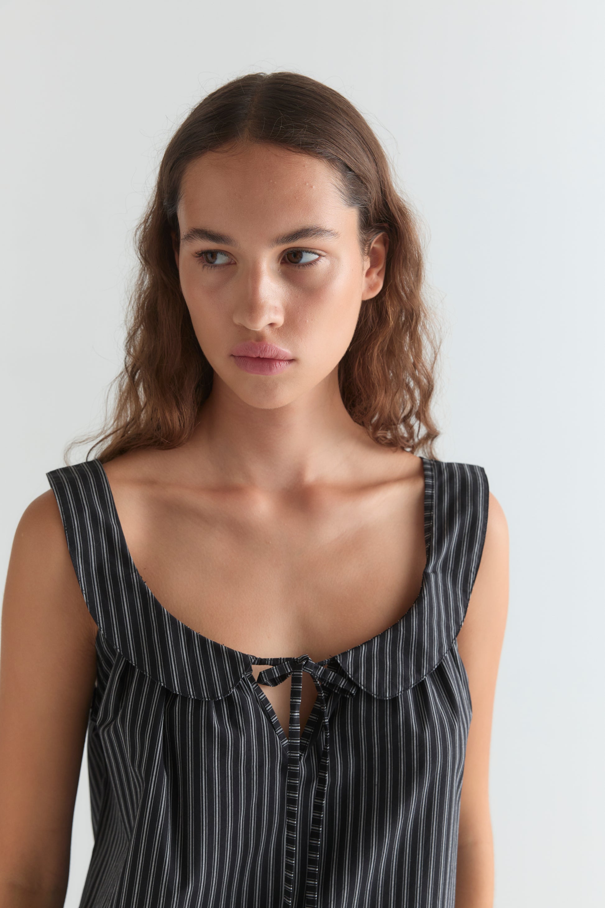 A woman with long, wavy brown hair wears the Deiji Studios Rounded Neck Dress - Mono stripe, a sleeveless black-and-white pinstripe dress with a rounded neckline and front bow. She gazes to the side with a neutral expression.
