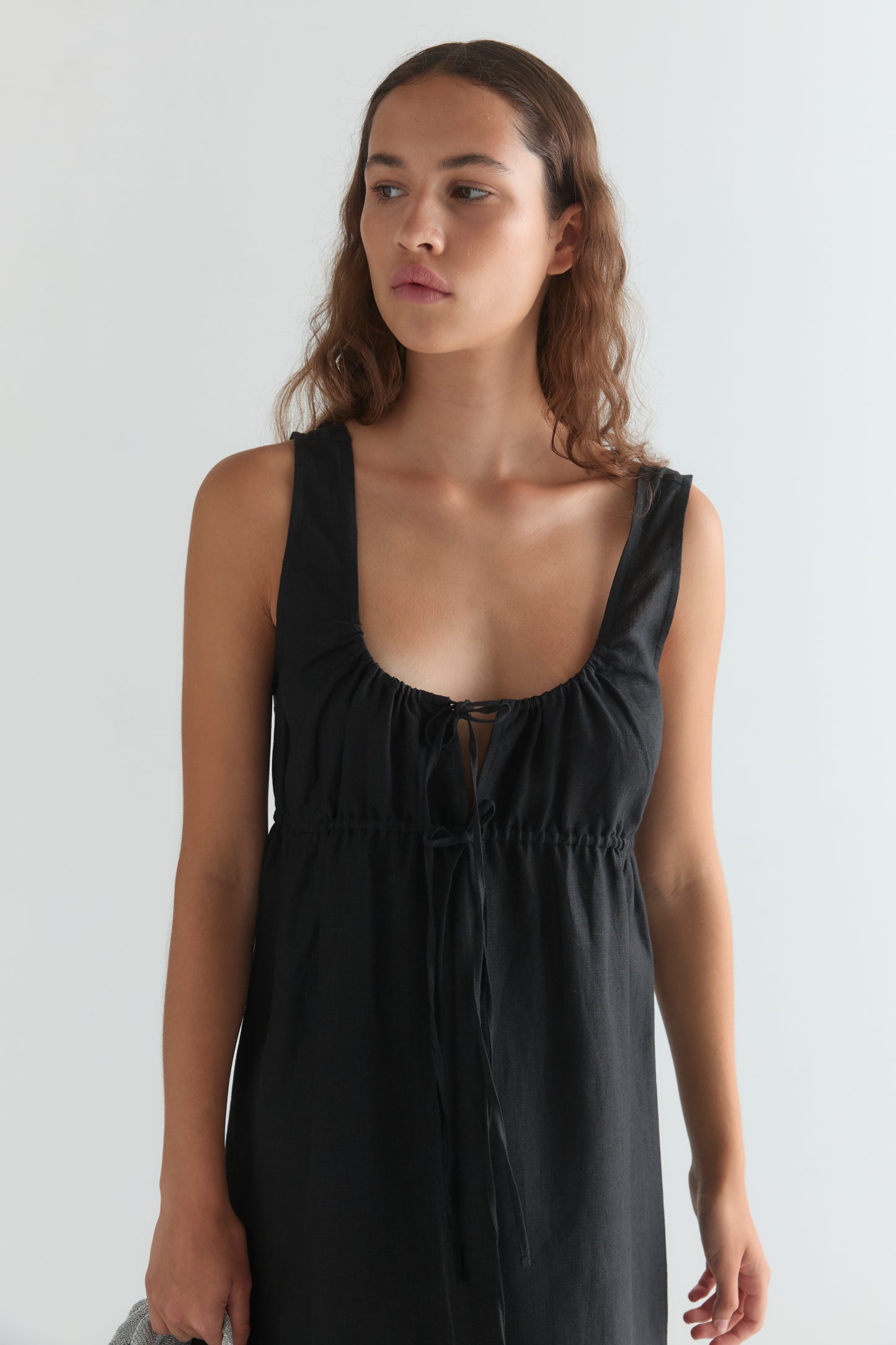 A woman with wavy brown hair wears a black Sleeveless Cord Dress featuring a low neckline and adjustable drawcord, standing against a plain light background, looking slightly to her left.