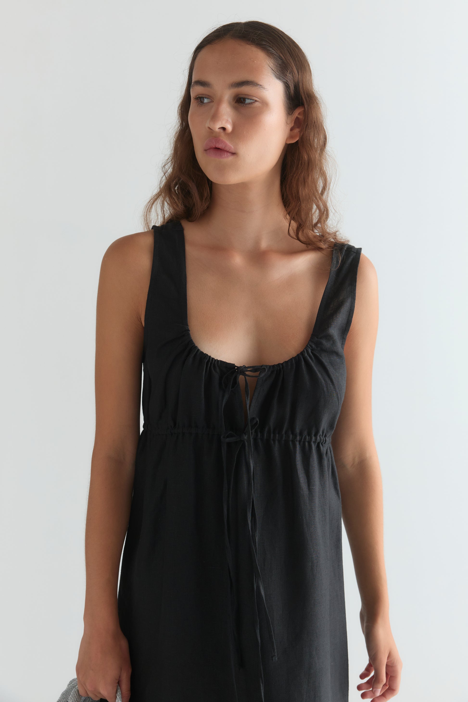 A woman with wavy brown hair wears a black Sleeveless Cord Dress featuring a low neckline and adjustable drawcord, standing against a plain light background, looking slightly to her left.