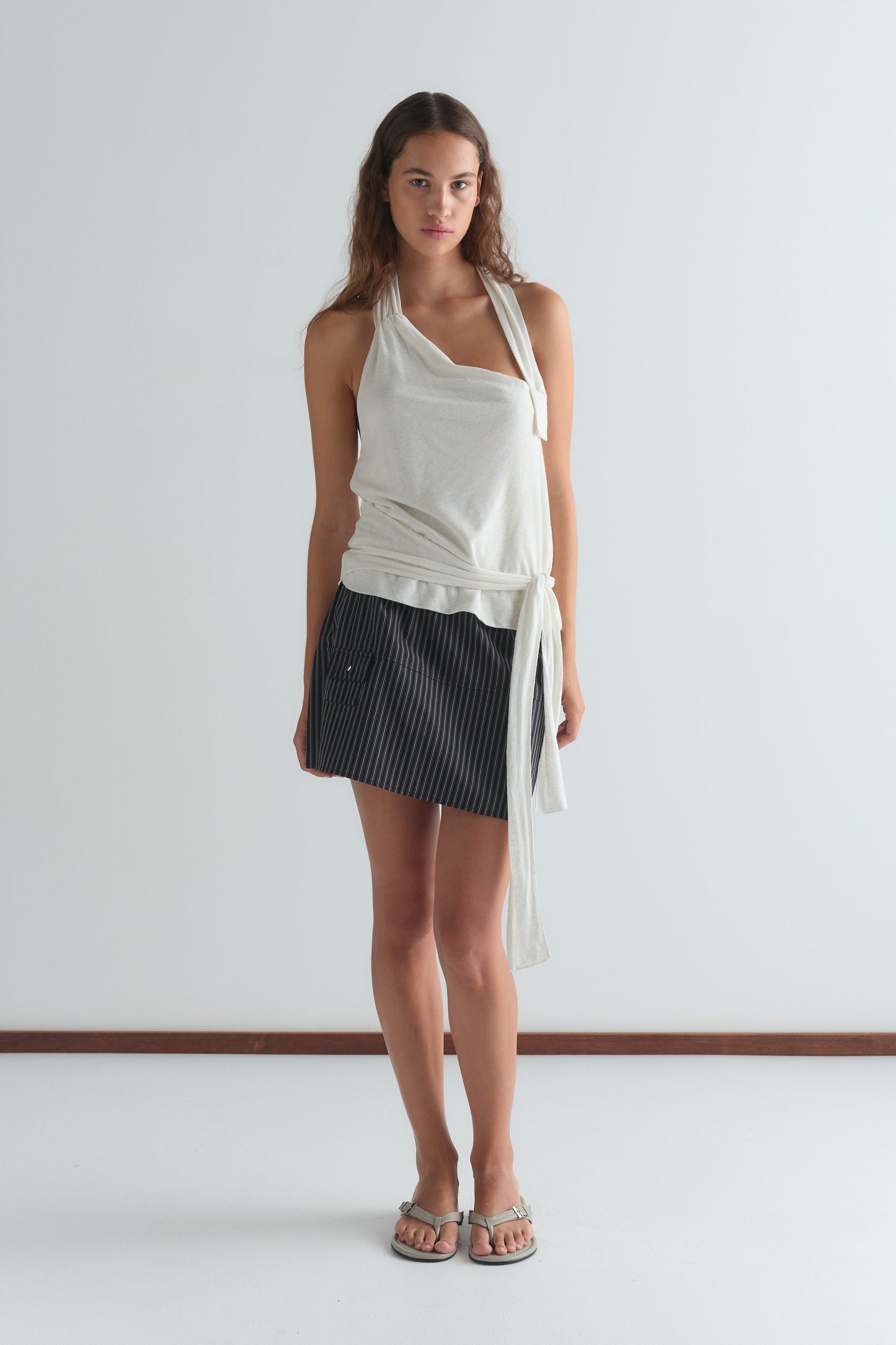 A woman stands against a light grey background wearing a cream asymmetric halter top, silver sandals, and the Pocket Skirt—a black pinstripe mini skirt.