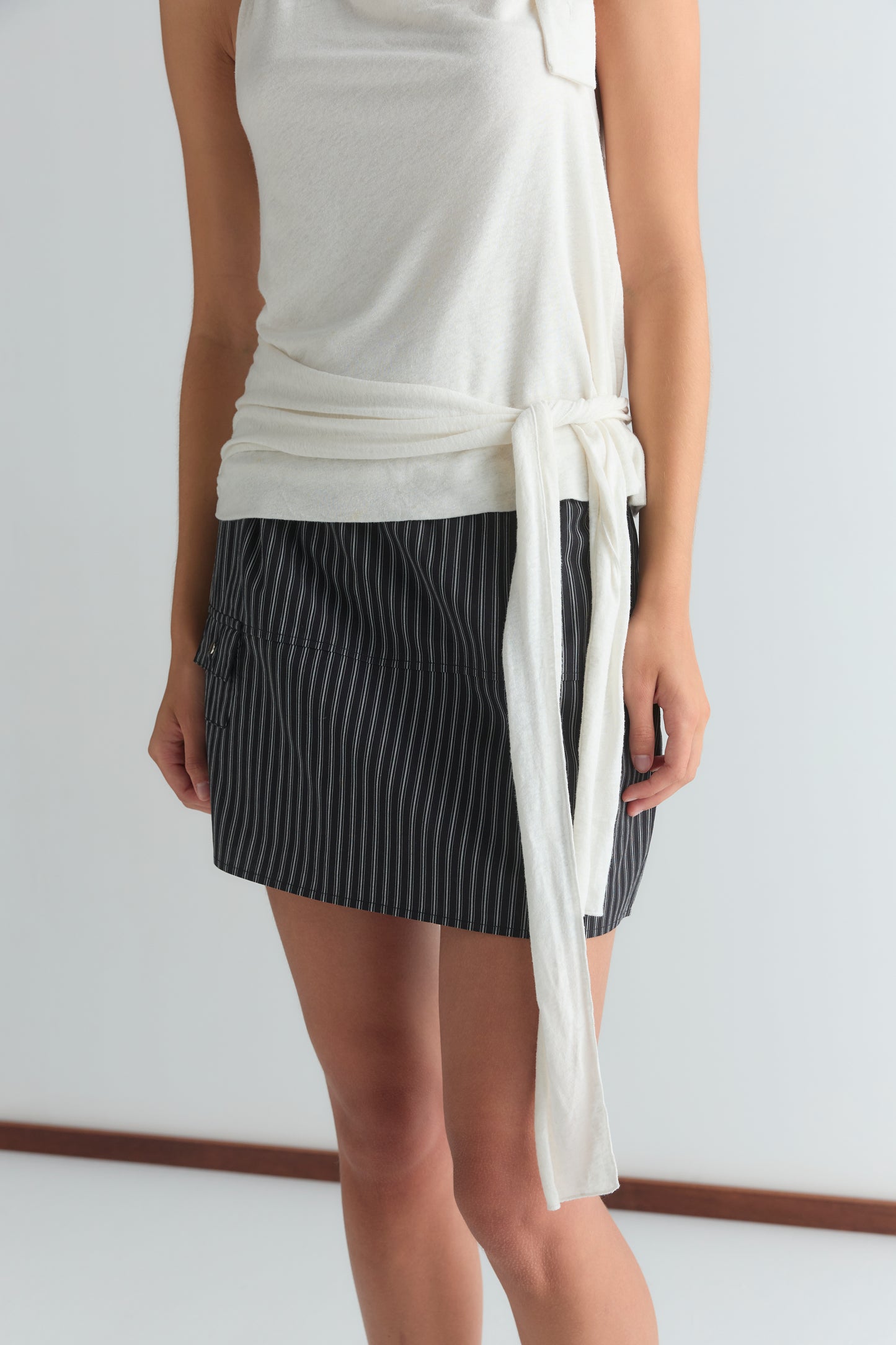 A person is wearing a sleeveless white wrap top with a side tie and the Pocket Skirt - Mono Stripe, a black pinstripe mini skirt, standing against a plain white background. The person's head is not visible in the image.