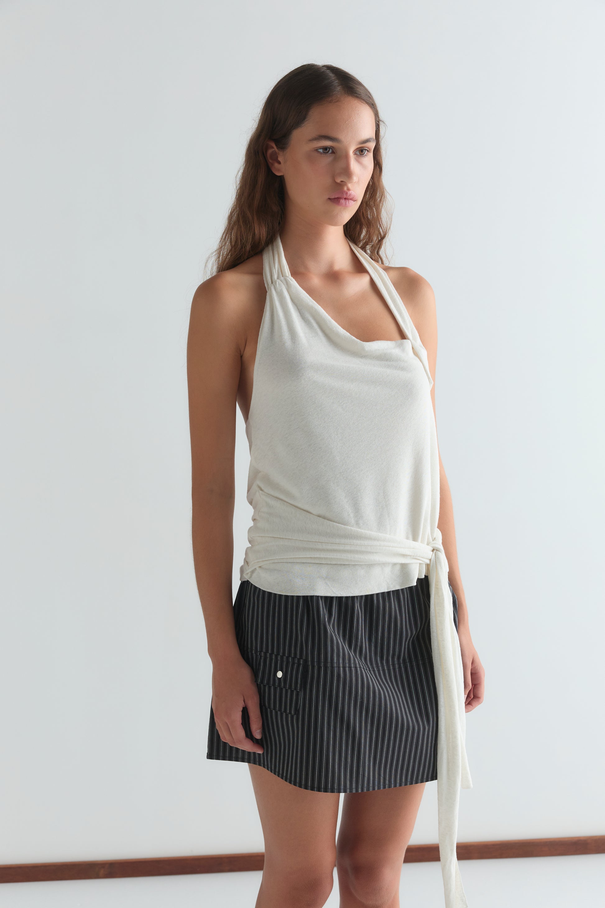 A woman with long brown hair and a neutral expression wears a sleeveless white halter top with a waist tie and the Pocket Skirt - Mono Stripe, standing against a plain white background.