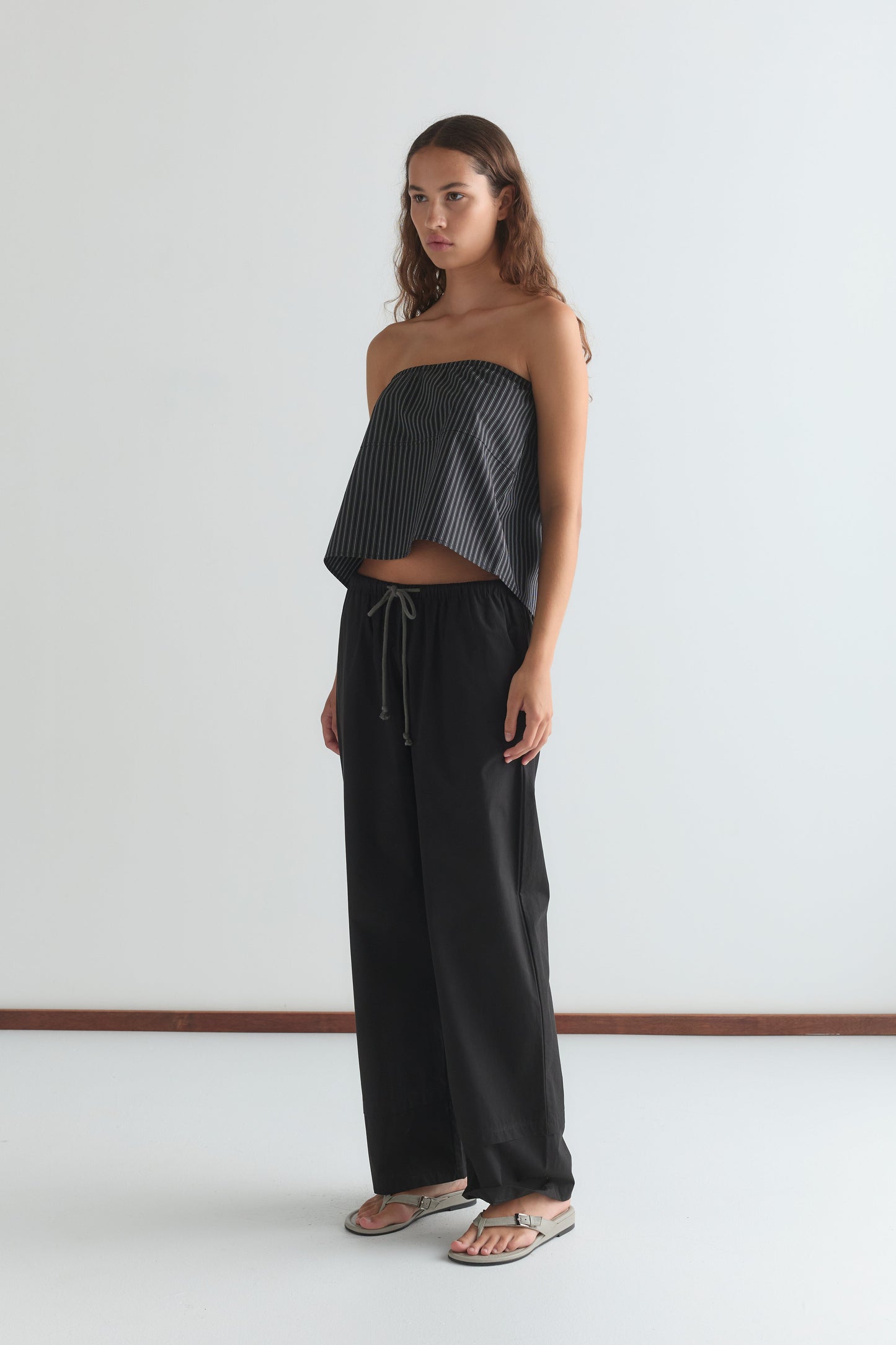 A woman with long, wavy brown hair and a neutral expression stands indoors against a white wall, wearing a strapless black and white striped top, the Layered Pant - Black from Deiji Studios, and silver flip-flops.