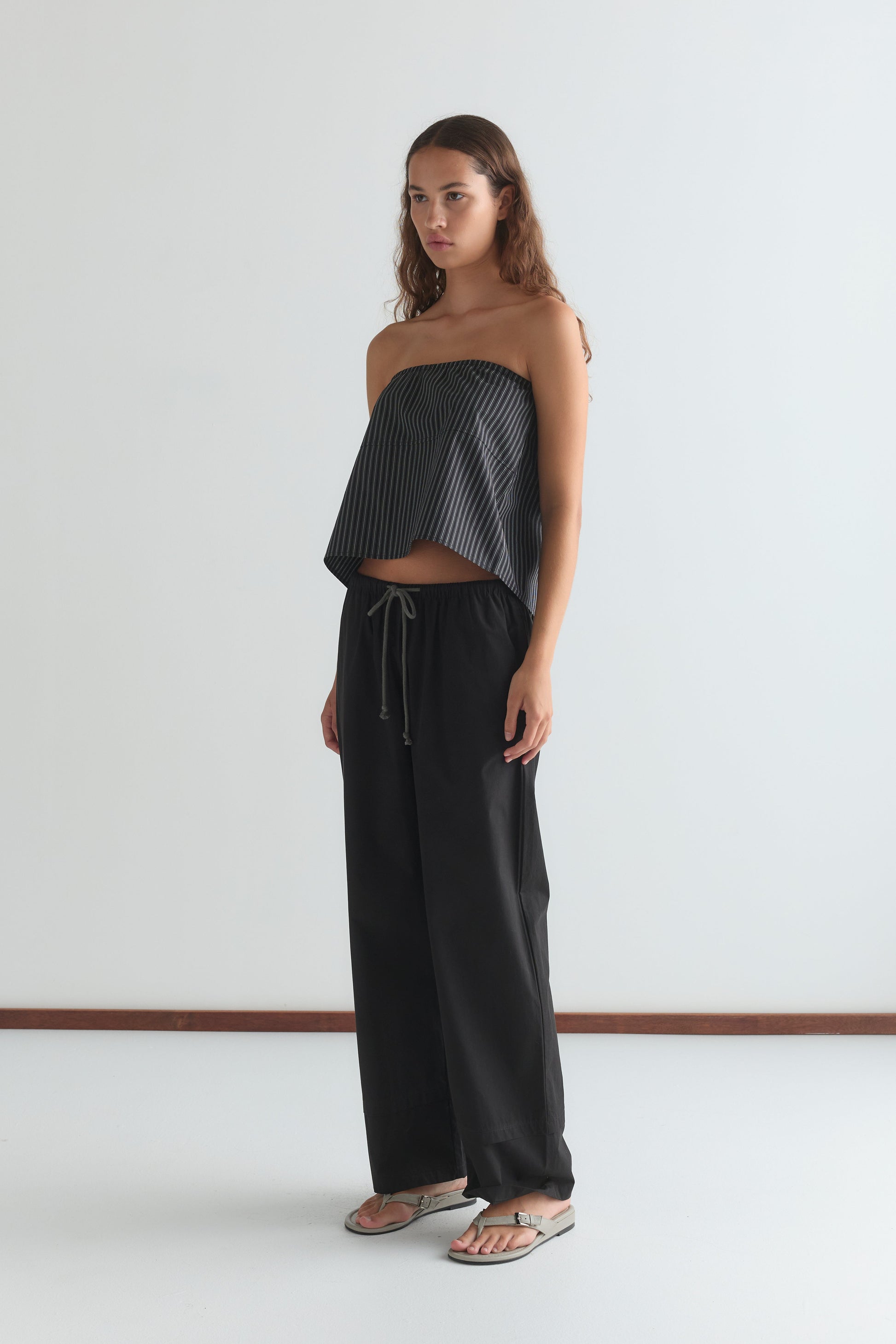 A woman with long, wavy brown hair and a neutral expression stands indoors against a white wall, wearing a strapless black and white striped top, the Layered Pant - Black from Deiji Studios, and silver flip-flops.