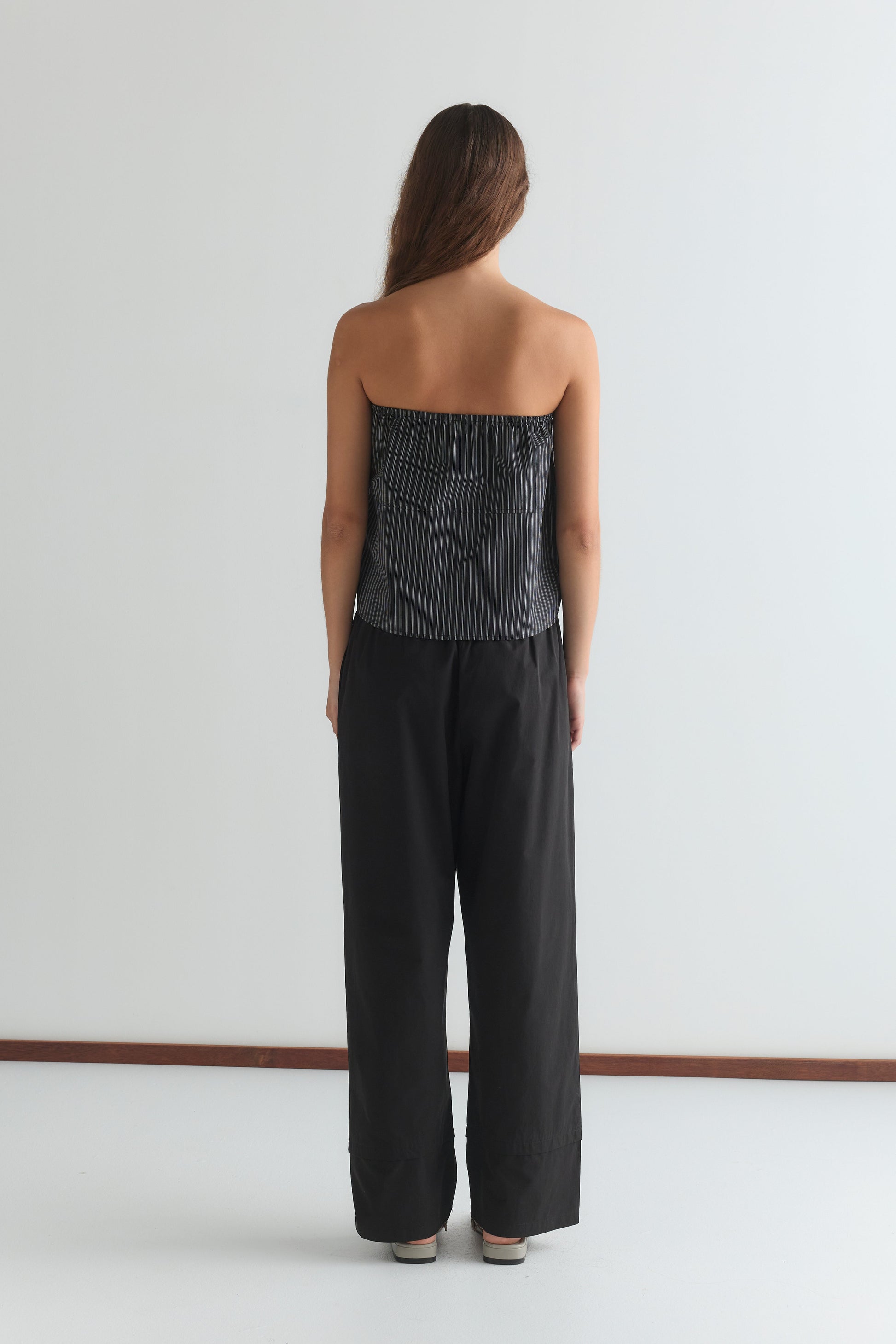 A person with long brown hair stands facing away, wearing a Deiji Studios black and white striped strapless top and the Layered Pant - Black against a plain white background.