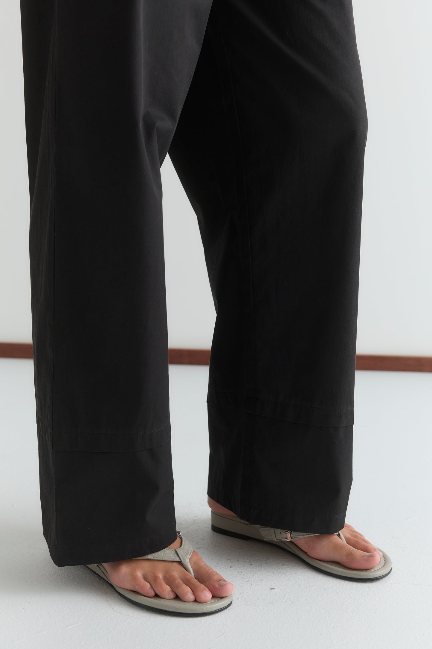 A person models the Layered Pant - Black by Deiji Studios, paired with light-colored flip-flops. Only their lower legs and feet are visible as they stand on a white floor against a plain backdrop.