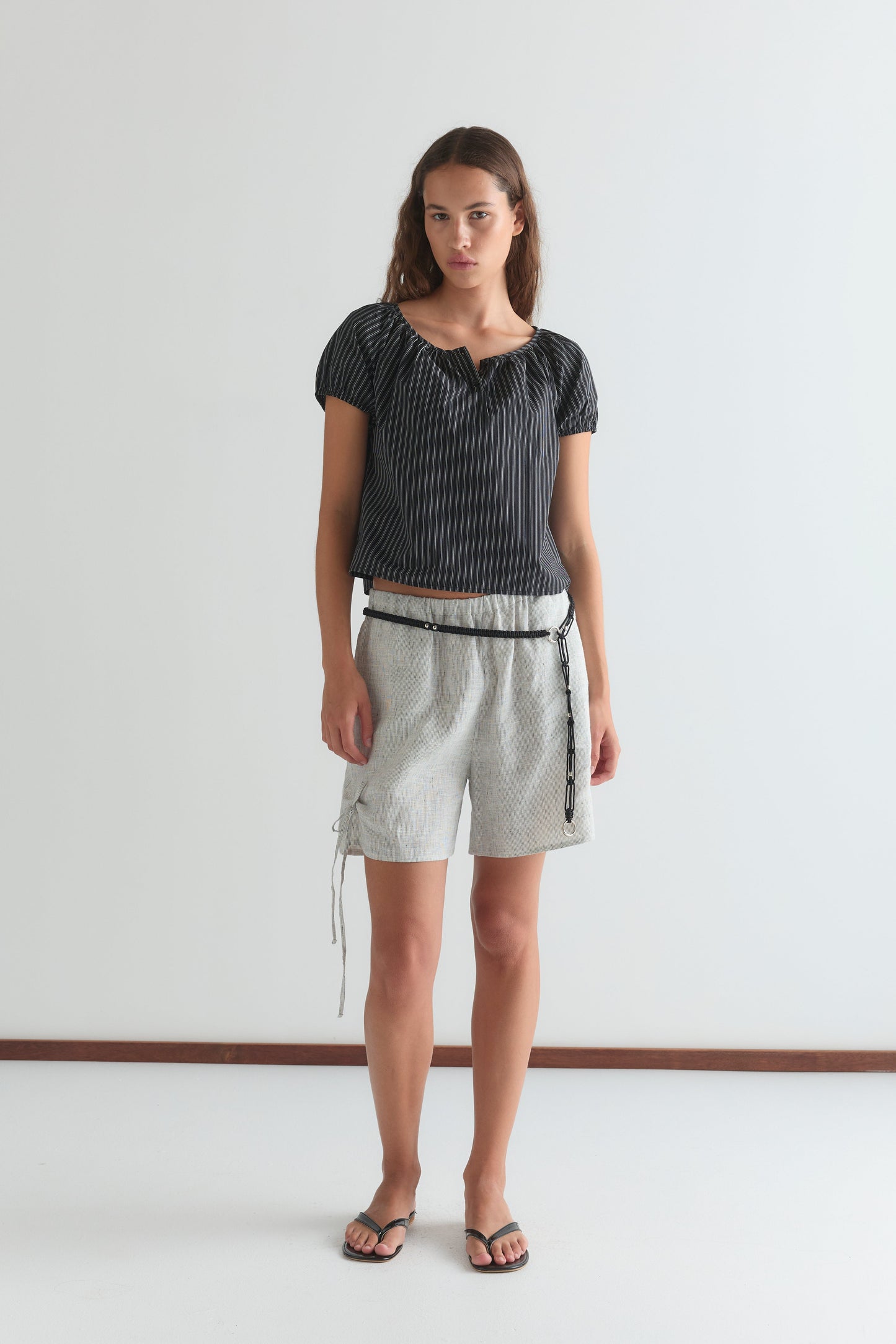 A woman stands against a plain white wall wearing the Poplin T Shirt - Mono Stripe by Deiji Studios, paired with light gray belted shorts and black flip-flops. She faces the camera with a neutral expression.