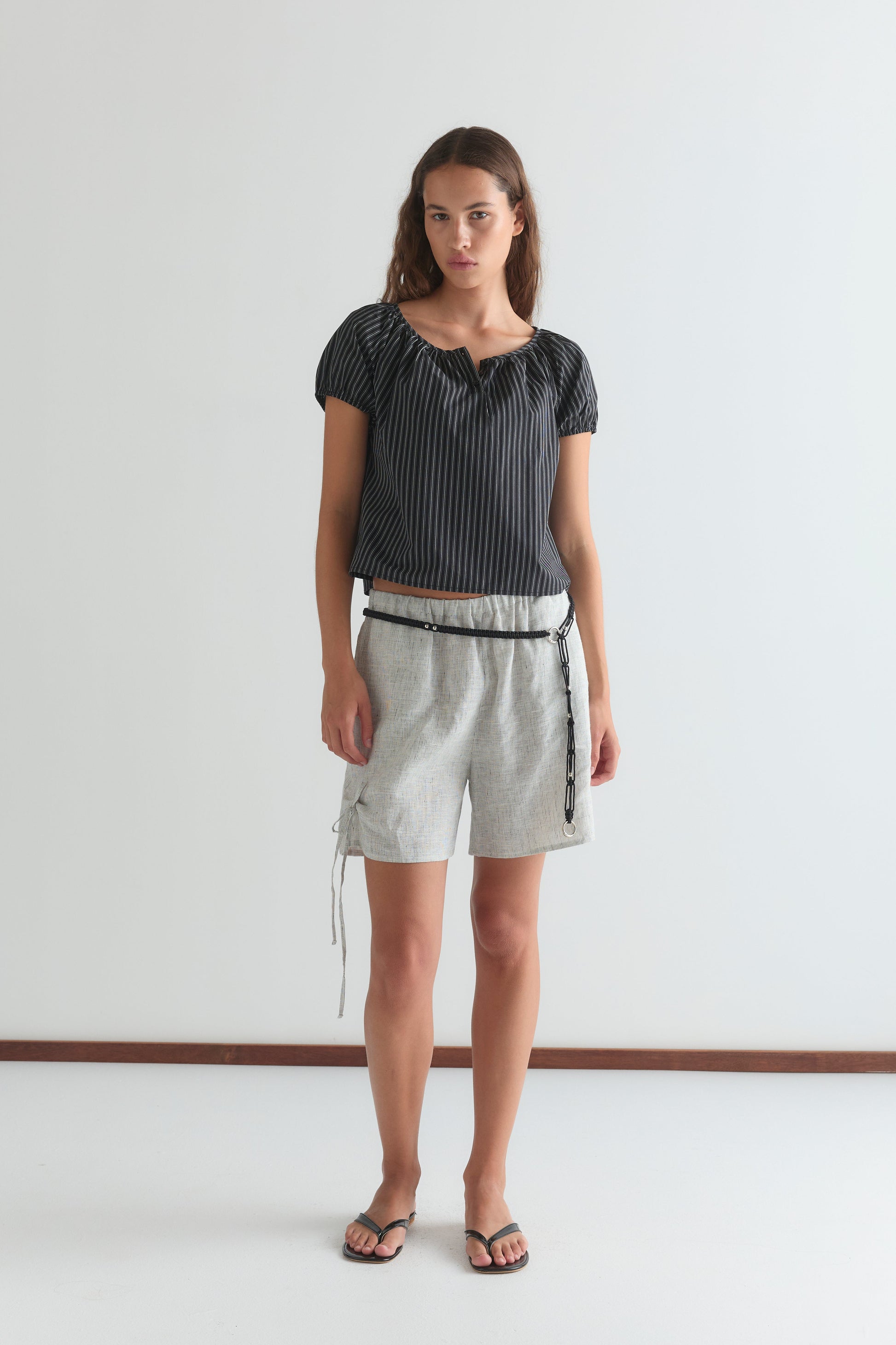 A woman stands against a plain white wall wearing the Poplin T Shirt - Mono Stripe by Deiji Studios, paired with light gray belted shorts and black flip-flops. She faces the camera with a neutral expression.