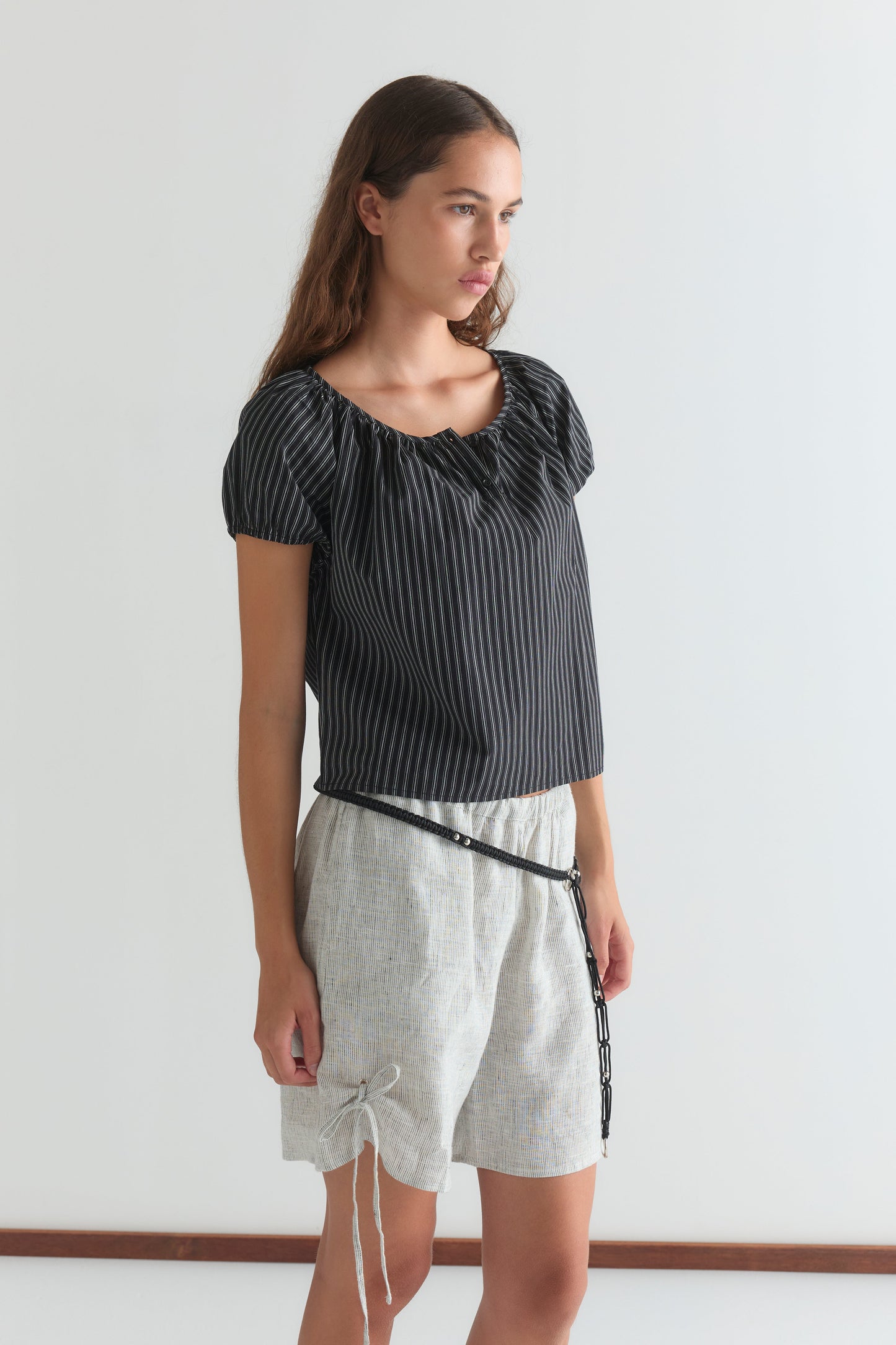 A woman with long brown hair stands against a white background, wearing a Deiji Studios Poplin T Shirt - Mono Stripe and light, loose shorts featuring a drawstring and thin black belt.