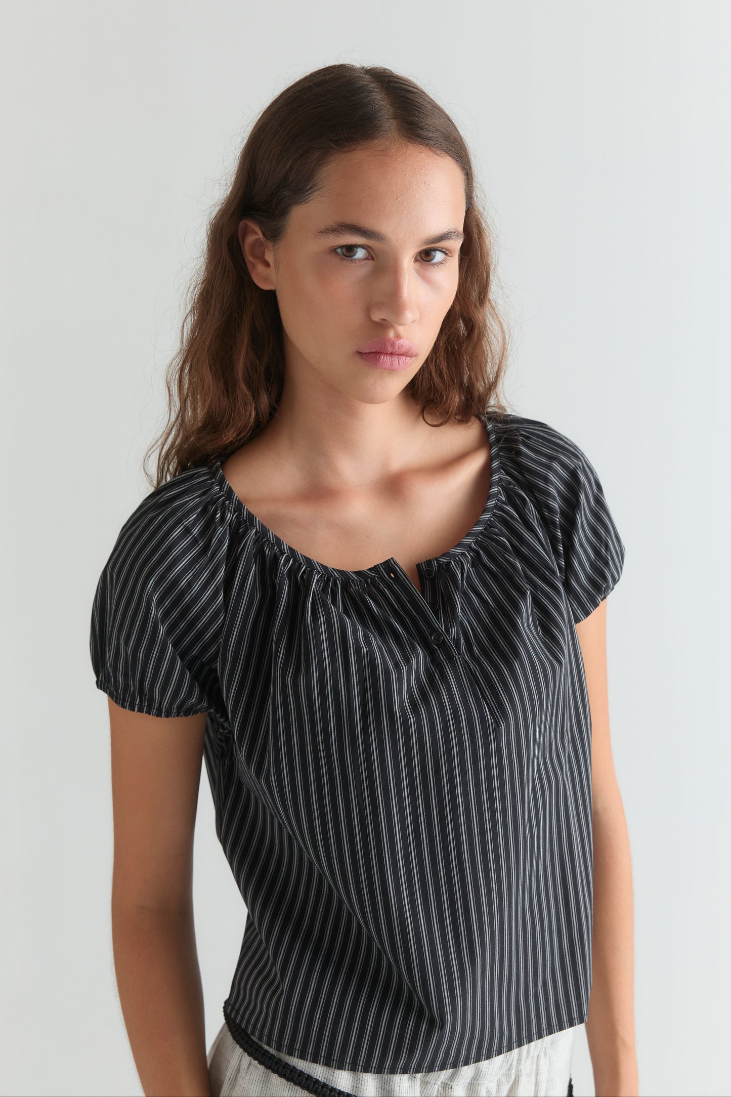 A young woman with long brown hair wears the Poplin T Shirt - Mono Stripe by Deiji Studios and light-colored pants, standing against a plain light background.