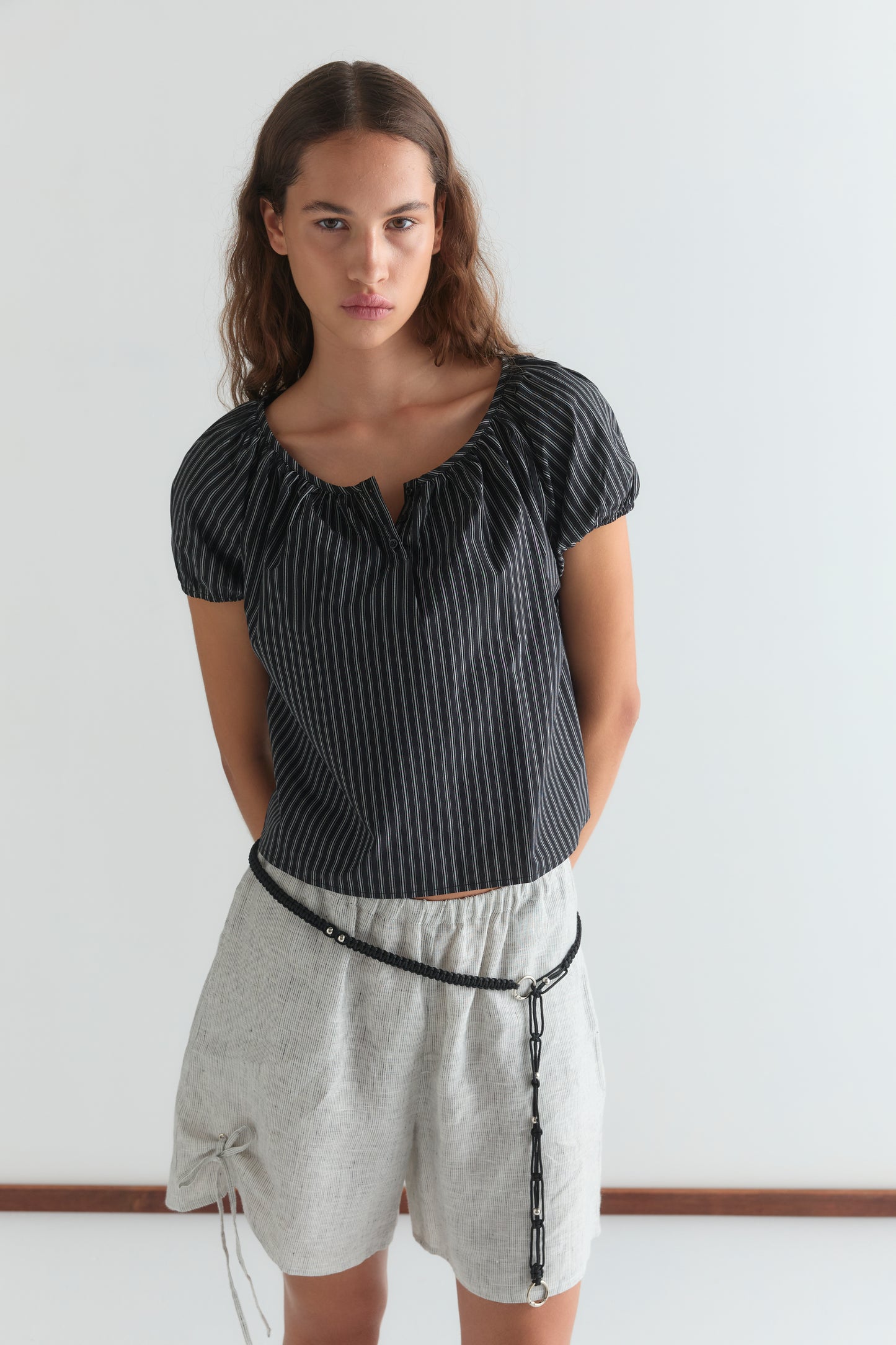 A woman stands against a white background, wearing the Poplin T Shirt - Mono Stripe from Deiji Studios with loose light gray shorts and a thin black belt low on her waist. Her hands are behind her back.
