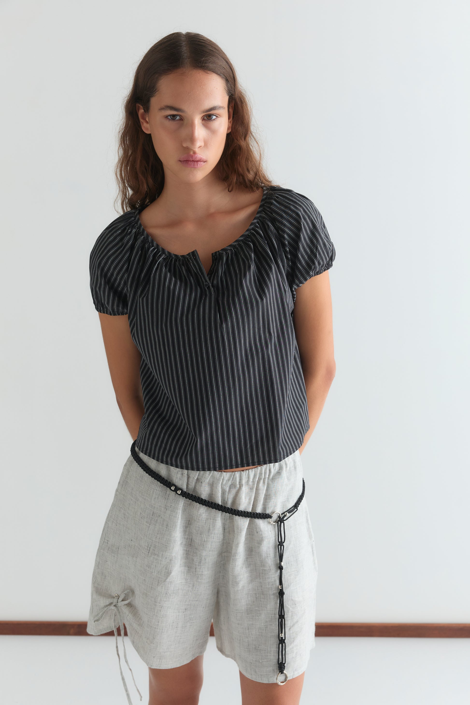 A woman stands against a white background, wearing the Poplin T Shirt - Mono Stripe from Deiji Studios with loose light gray shorts and a thin black belt low on her waist. Her hands are behind her back.