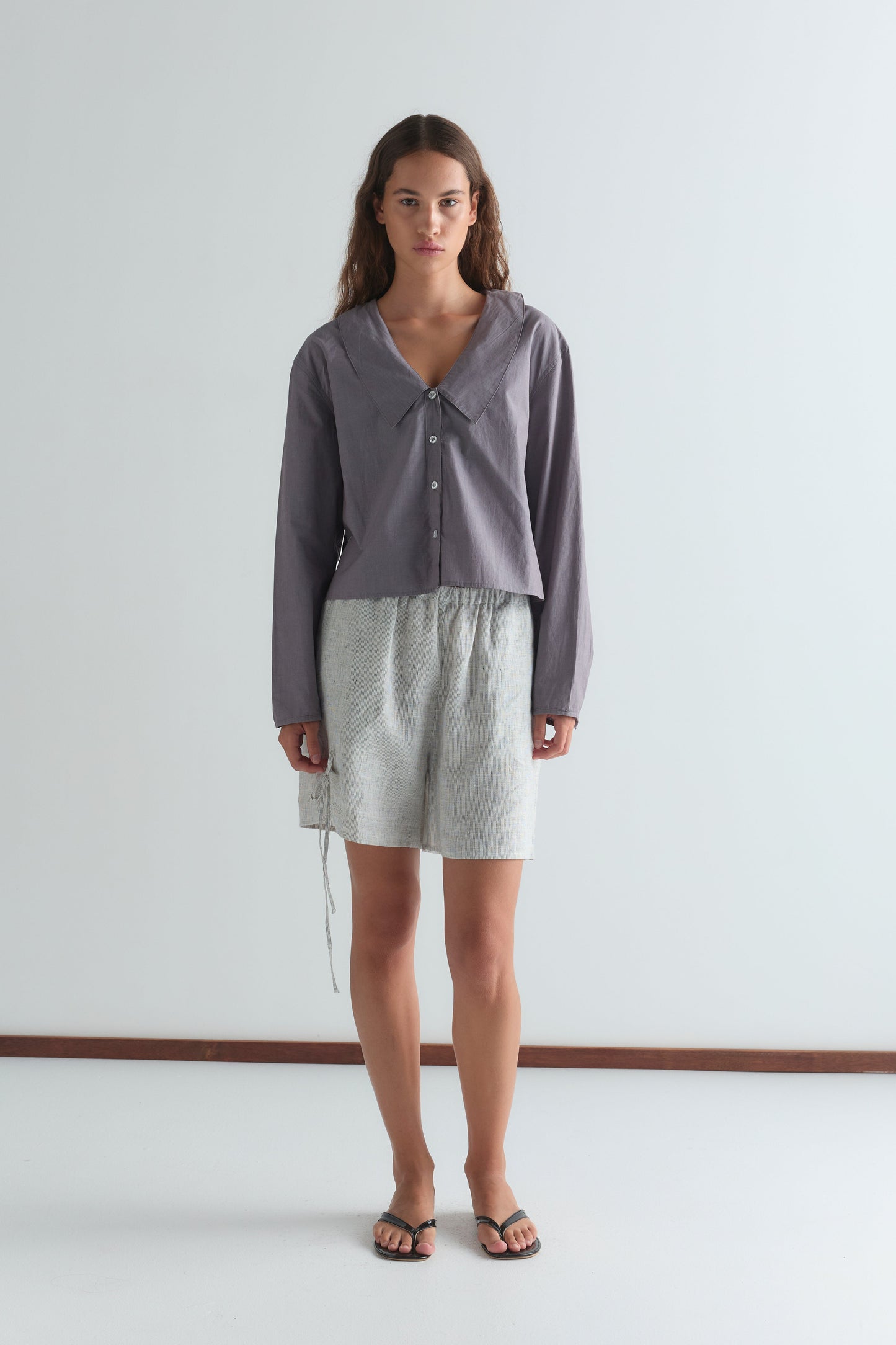 A woman stands indoors against a plain white wall, wearing a loose gray blouse from Deiji Studios, the Eyelet Short - Fine Stripe in light gray with a drawstring waist, and black flip-flops. She has long brown hair and a neutral expression.