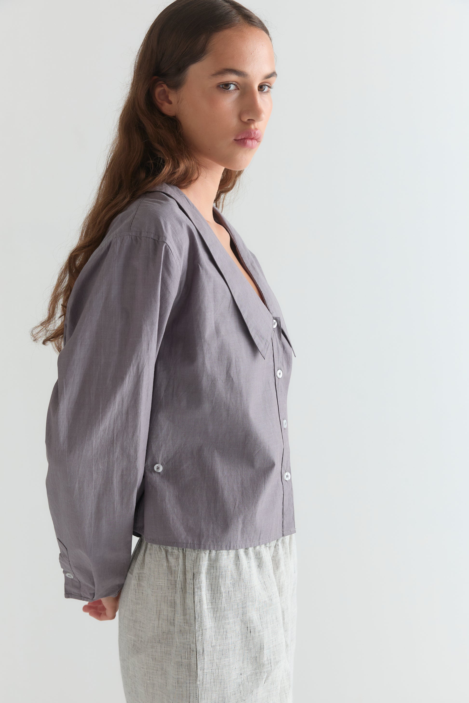 A woman with long brown hair wears the Deiji Studios Waist Button Shirt in Currant and light-colored pants, standing sideways against a plain light background, looking toward the camera with a neutral expression.
