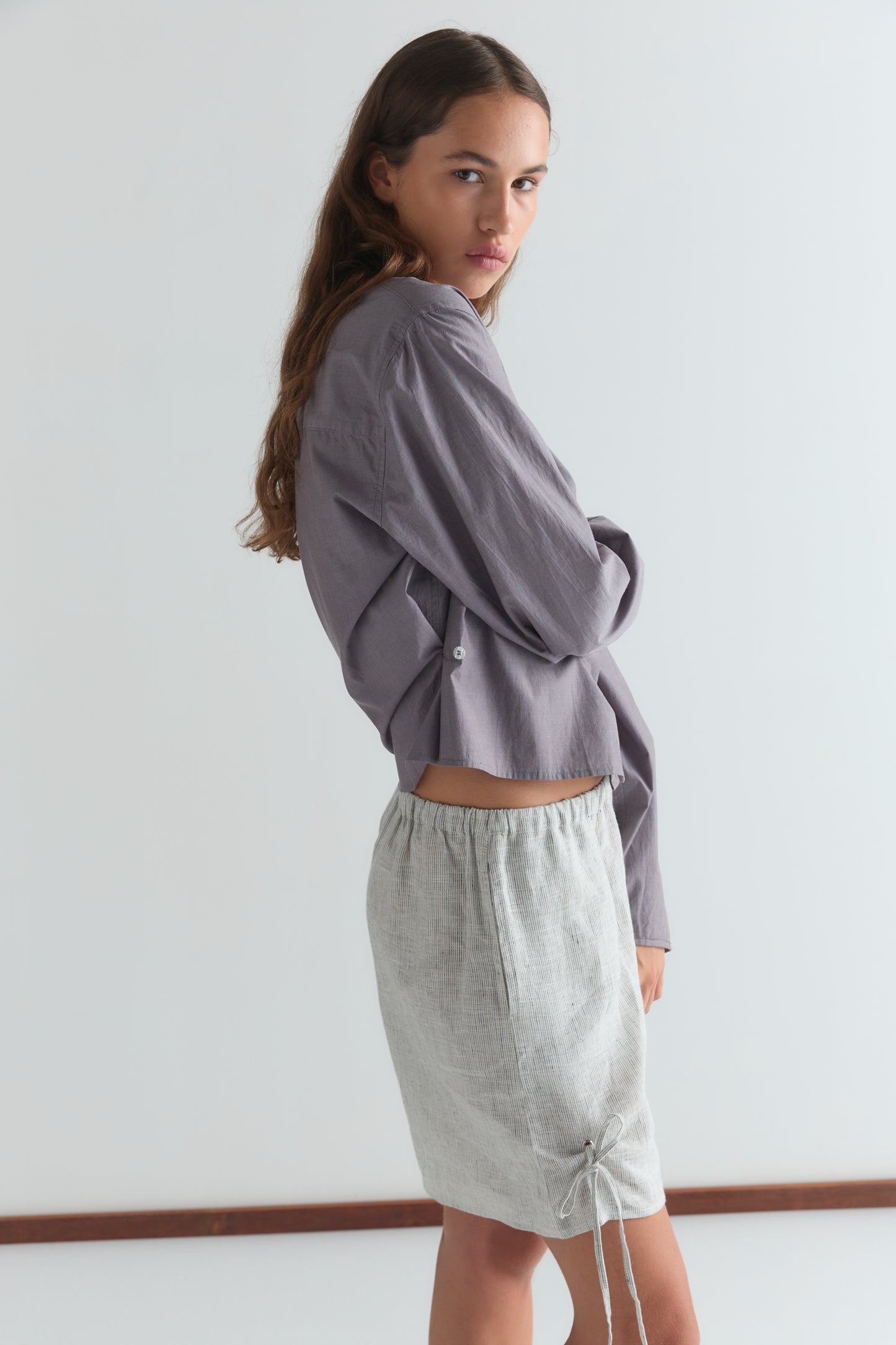 A woman with long brown hair wears a loose, long-sleeved lavender shirt and Fine Stripe Eyelet Shorts, standing sideways and looking over her shoulder against a plain white background.