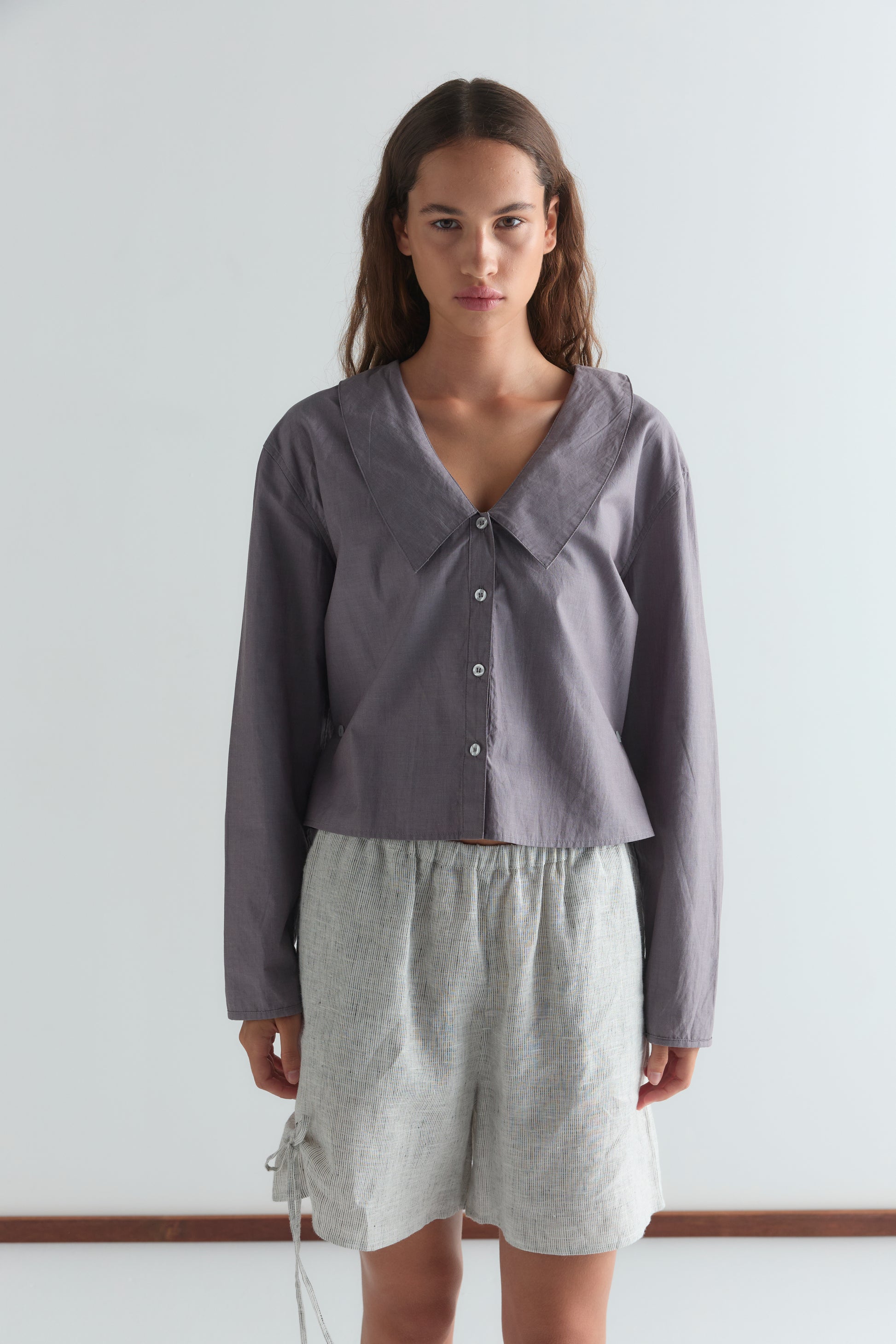 A woman with long brown hair and a neutral expression stands against a plain white background, wearing the Waist Button Shirt - Currant by Deiji Studios and light gray textured shorts.