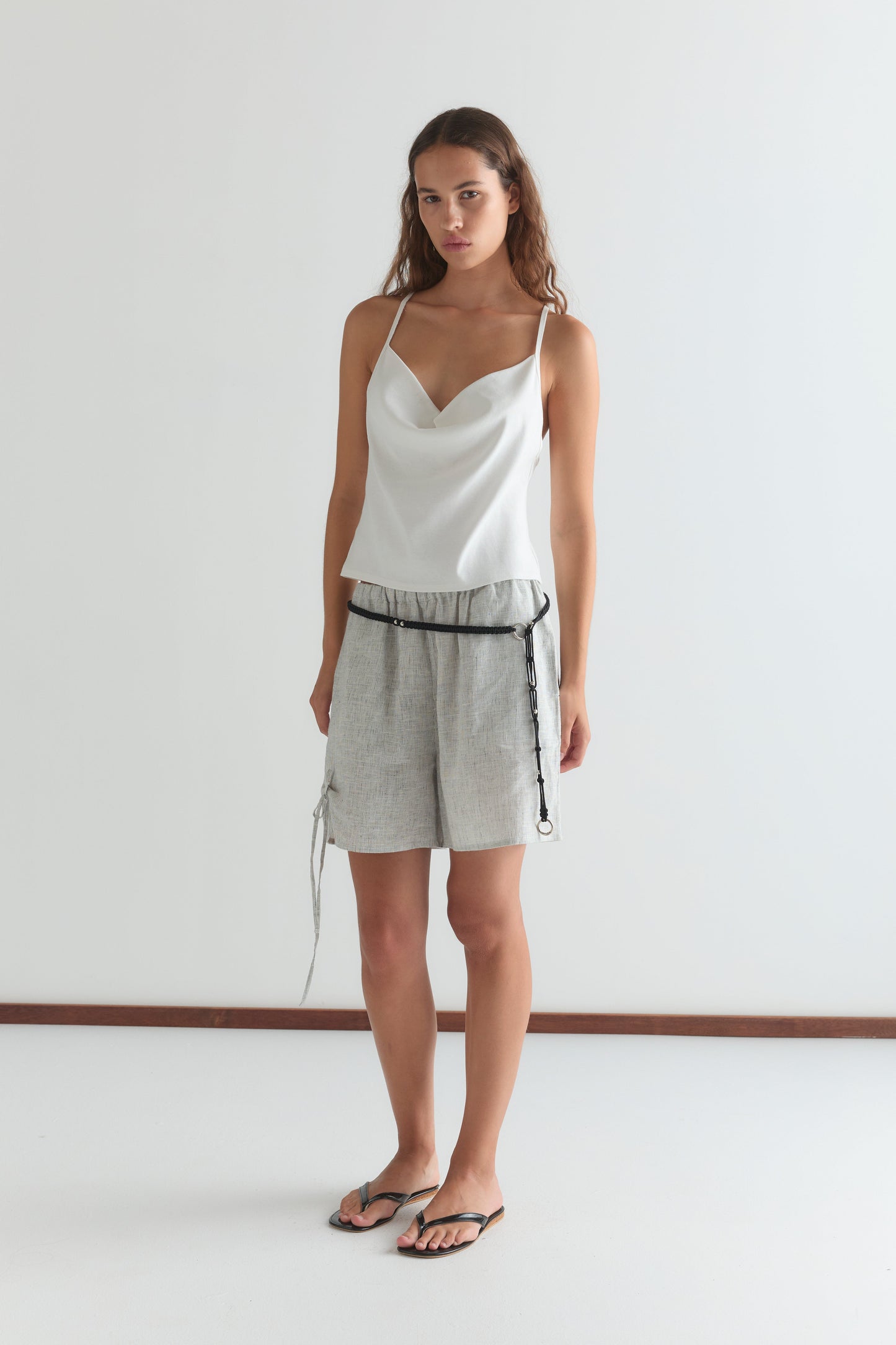 A woman stands against a white wall wearing a white sleeveless top, Deiji Studios Eyelet Short - Fine Stripe with a black tie belt, and black flip-flops. She has long brown hair and a neutral expression.