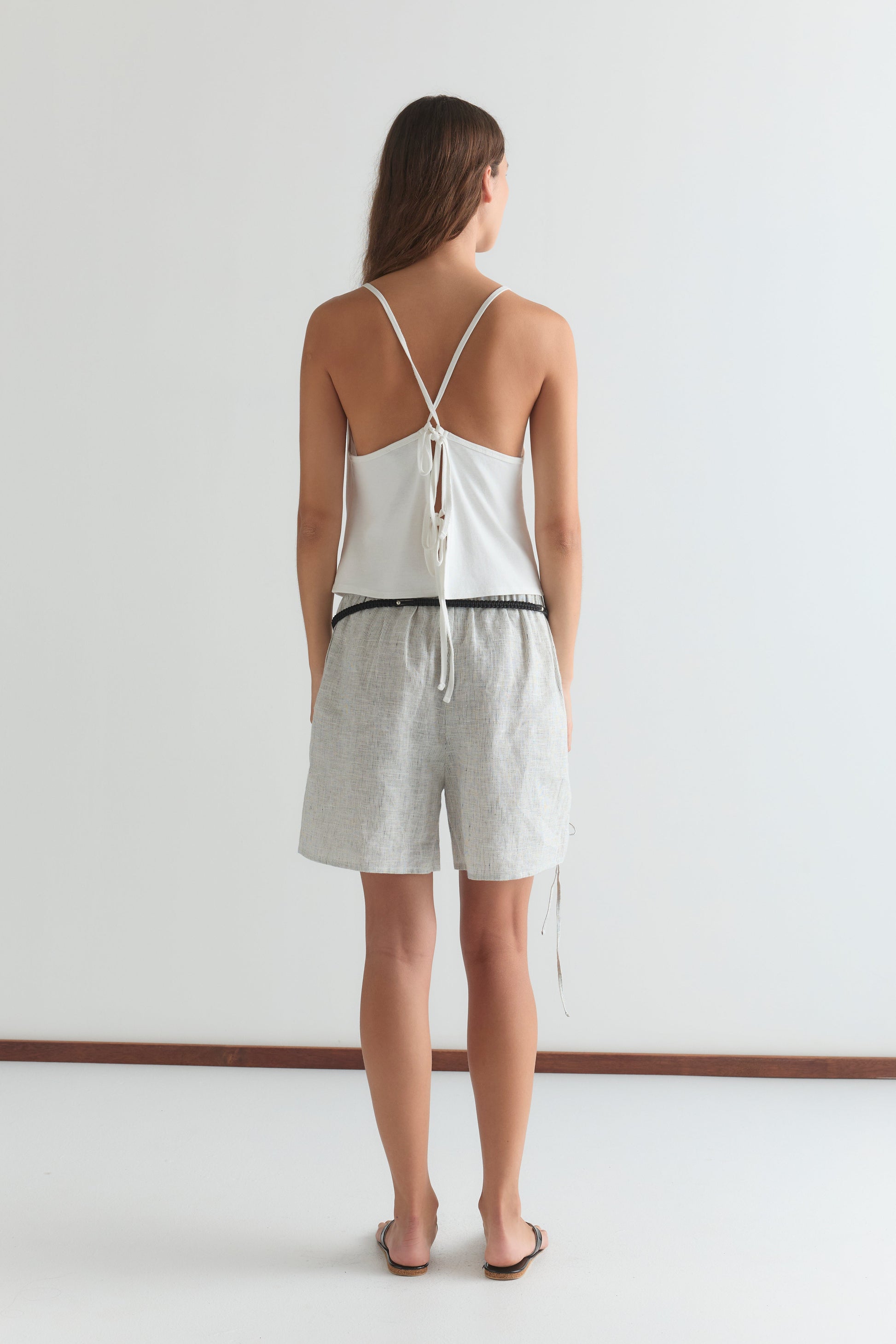 A woman with long brown hair wears the Deiji Studios Drape Jersey Top in white, featuring a back tie, paired with light-patterned shorts and sandals. She is facing away against a plain white background.