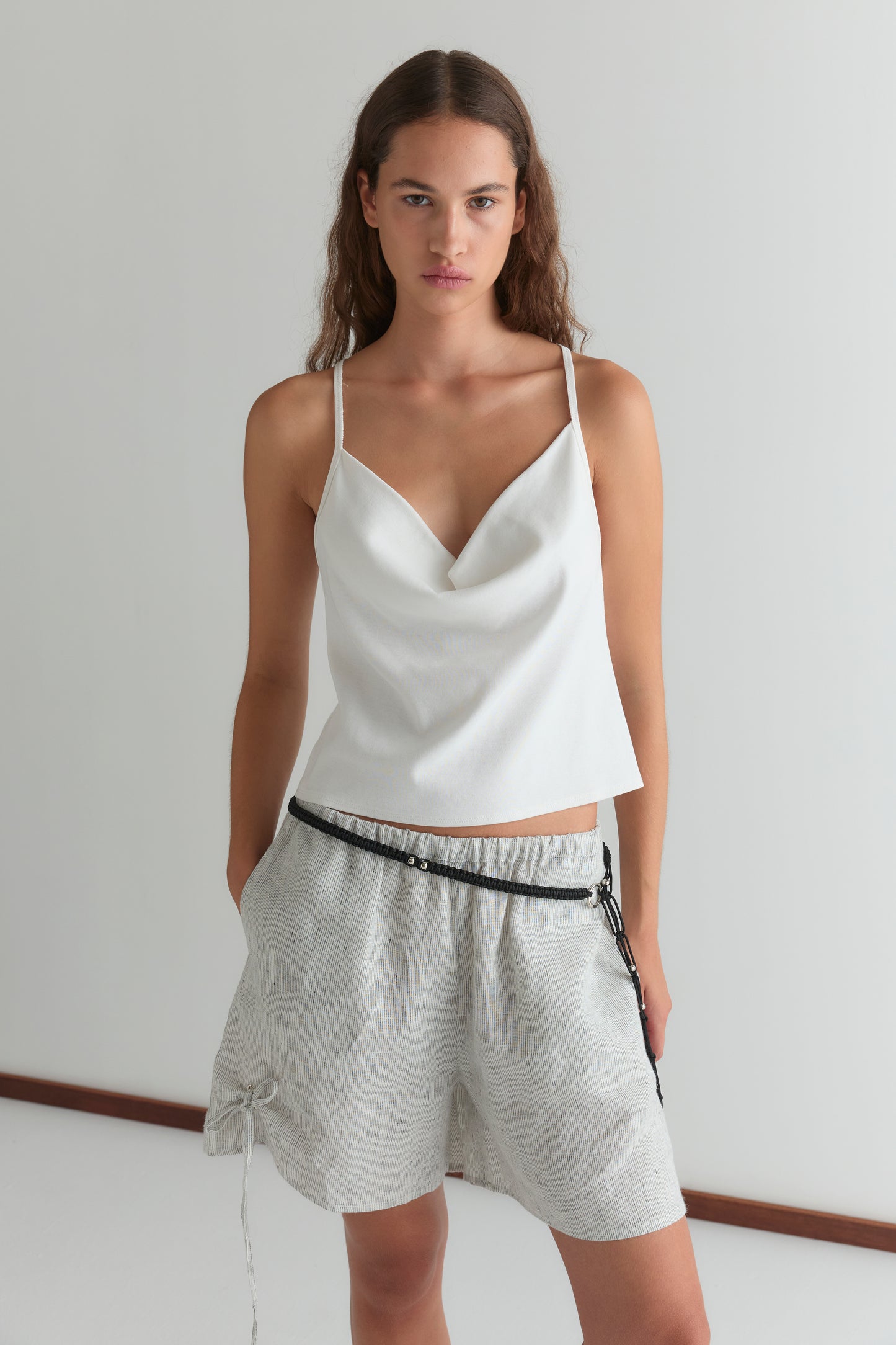 A woman with long wavy brown hair and a neutral expression stands against a plain white wall, wearing a white draped camisole and the Eyelet Short - Fine Stripe with a black belt.