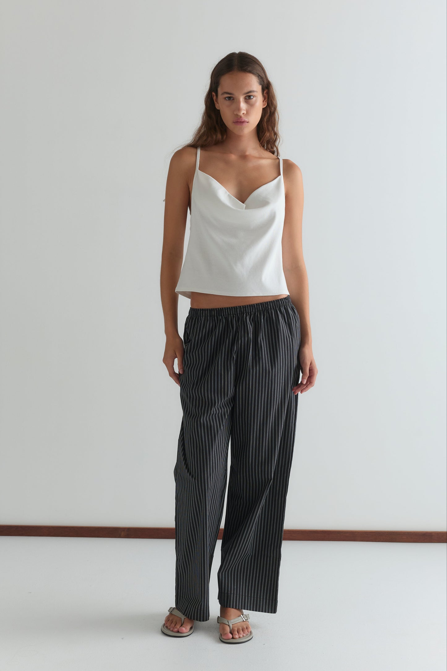 A woman with long brown hair and a neutral expression stands against a white wall wearing a white sleeveless cowl-neck top, sandals, and the Deiji Studios Ease Trouser - Mono Stripe.
