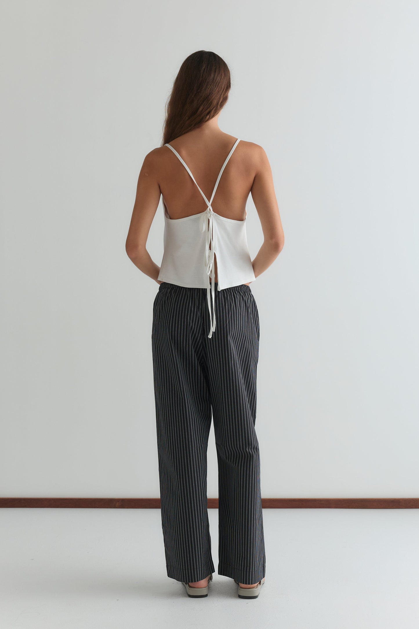 A person faces away from the camera, wearing a white backless tie-strap top and Deiji Studios' Ease Trouser - Mono Stripe, which is dark with pinstripes and ankle length. The background is simple and minimalistic.