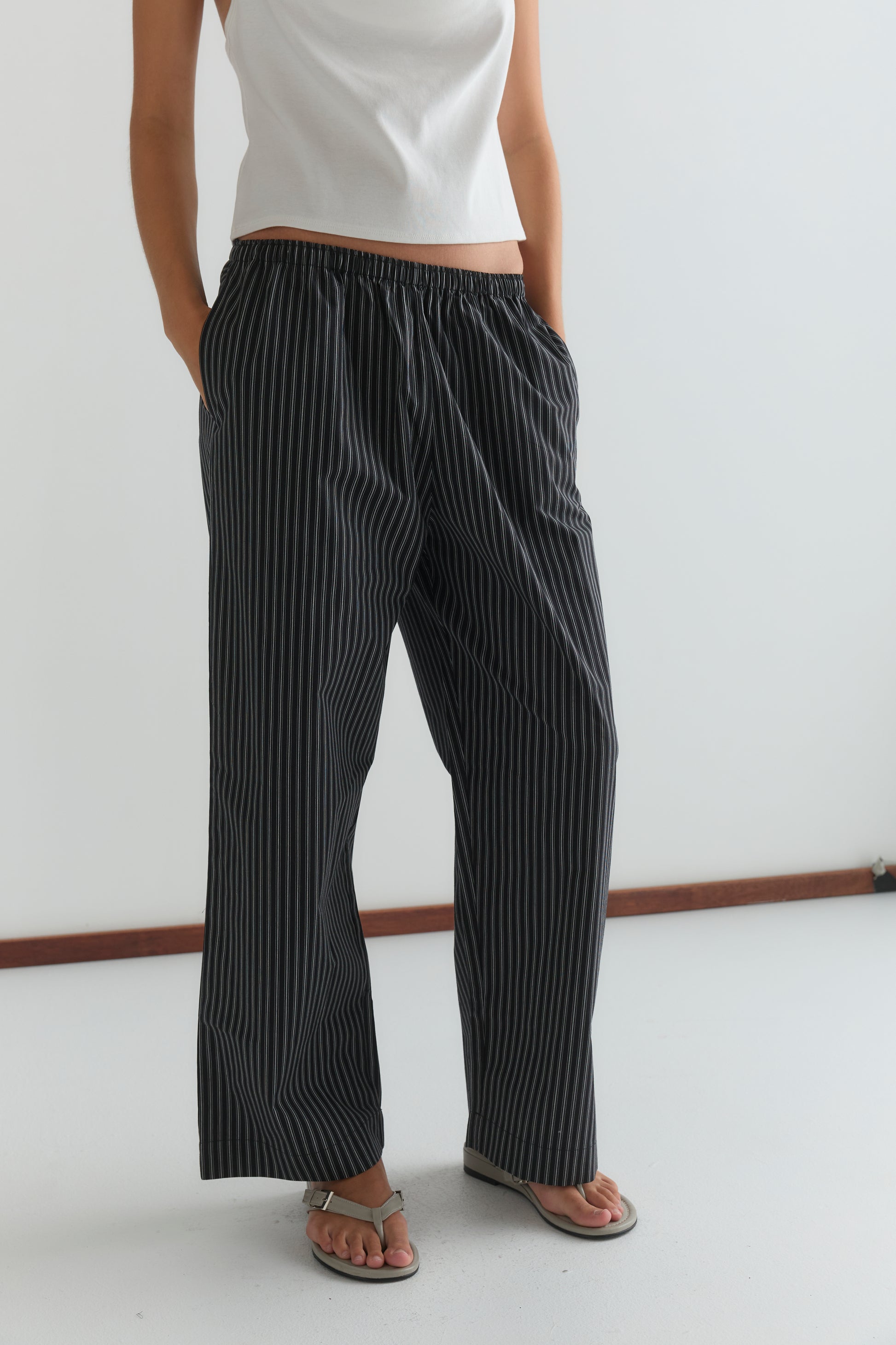 A person wearing a white sleeveless top, Ease Trouser - Mono Stripe by Deiji Studios, and sandals stands with hands in their pockets against a plain white background.