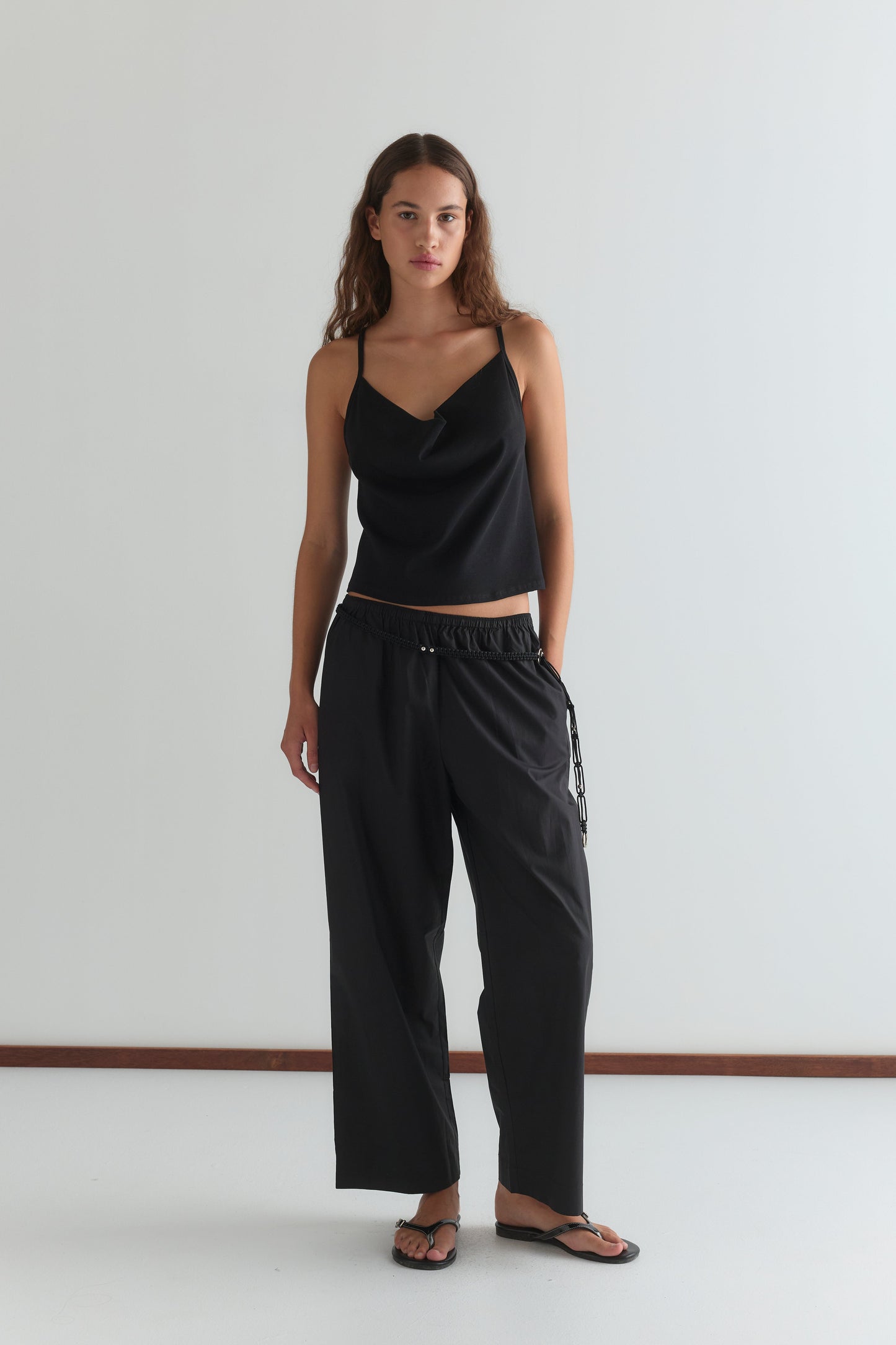 A woman stands against a plain white wall, wearing the Drape Jersey Top - Black from Deiji Studios, loose black drawstring pants, and black sandals. She has long wavy brown hair and a neutral expression.