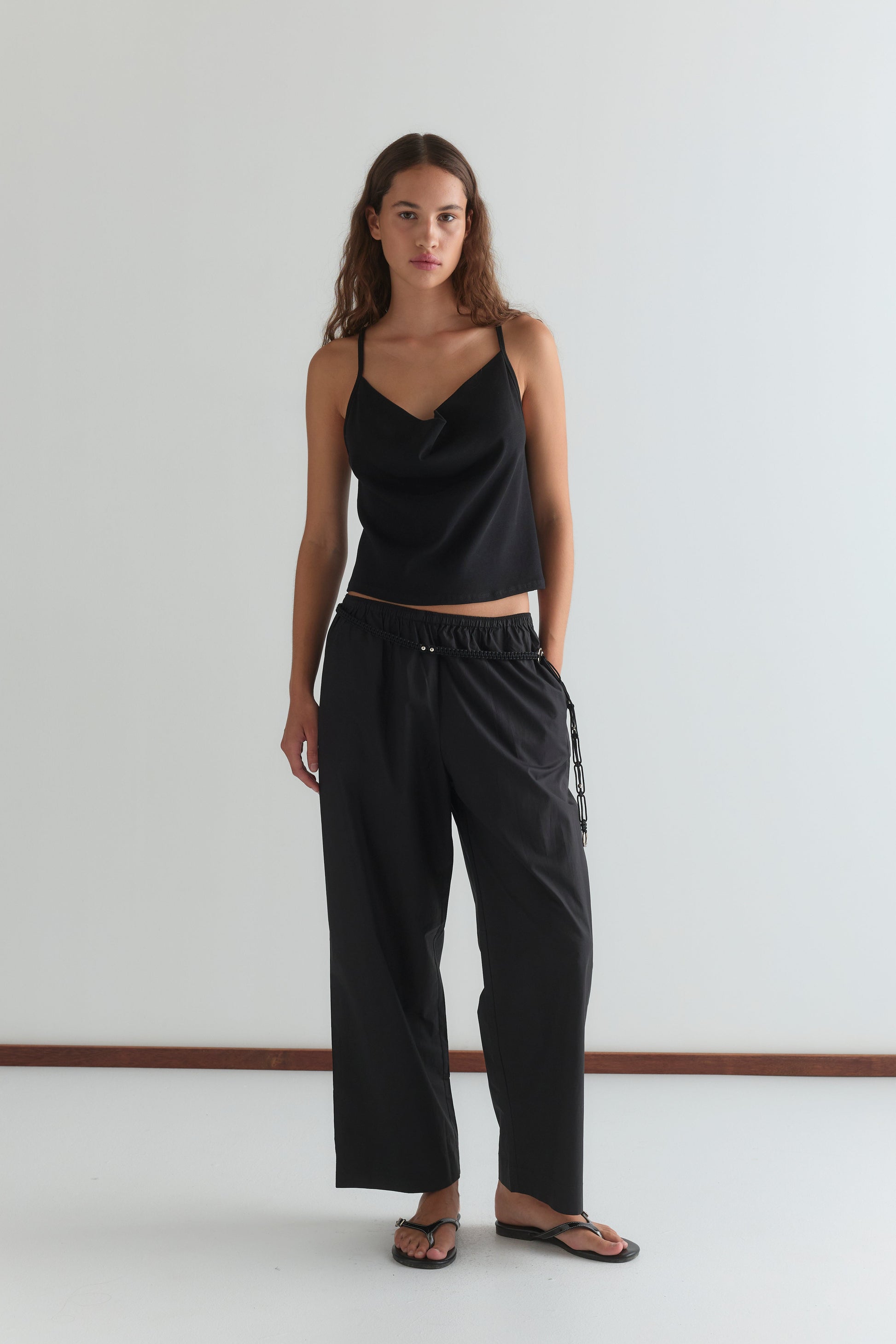 A woman stands against a plain white wall, wearing the Drape Jersey Top - Black from Deiji Studios, loose black drawstring pants, and black sandals. She has long wavy brown hair and a neutral expression.