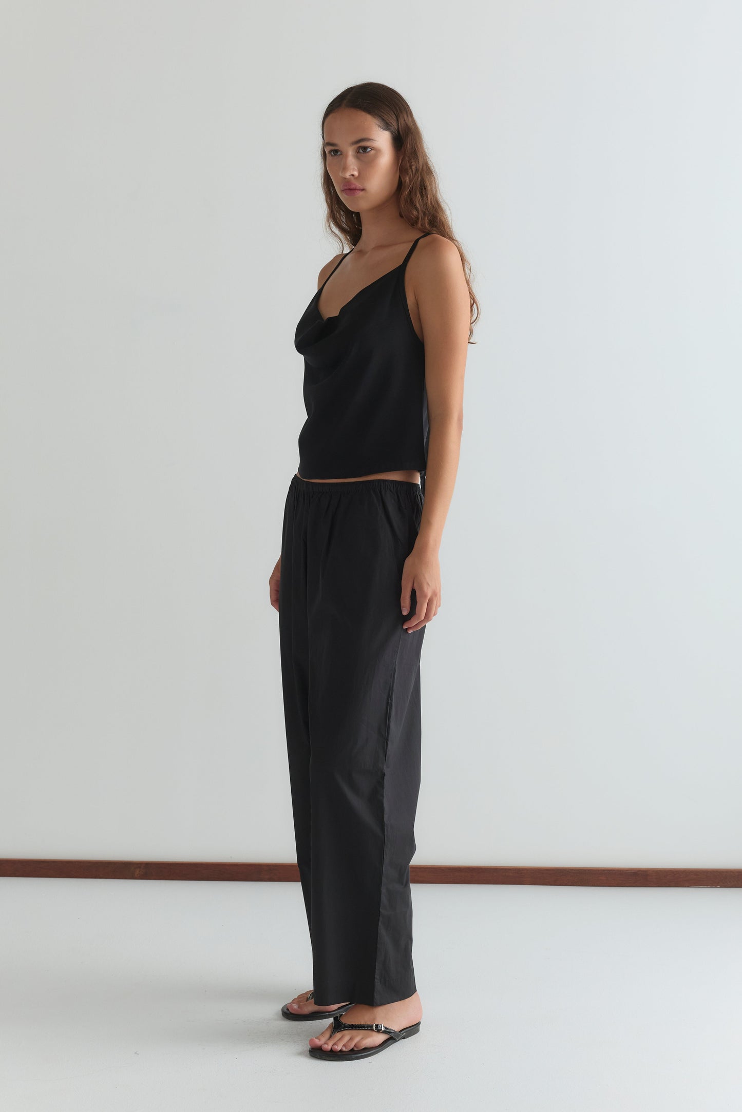A woman with long brown hair wears the Drape Jersey Top - Black by Deiji Studios, paired with black pants and sandals, standing against a plain white wall and looking slightly to the side with a neutral expression.
